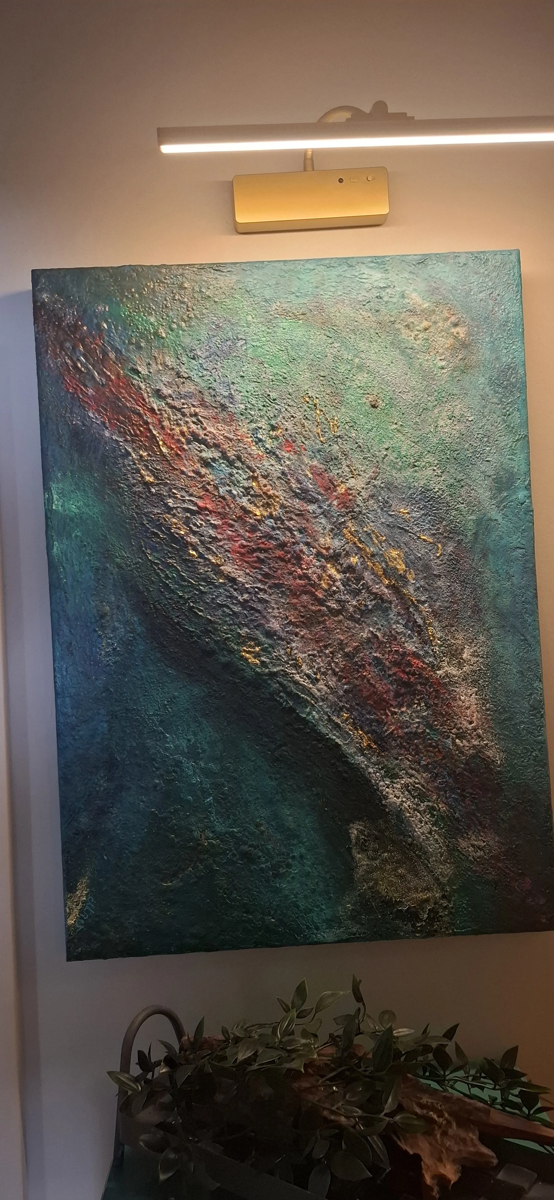 Abstraktes Gemälde mit Textur in Blau, Grün, Rot und Goldtönen, an der Wand mit LED-Lichtleiste darüber, darunter ein Pflanzgefäß mit grünen Blättern.