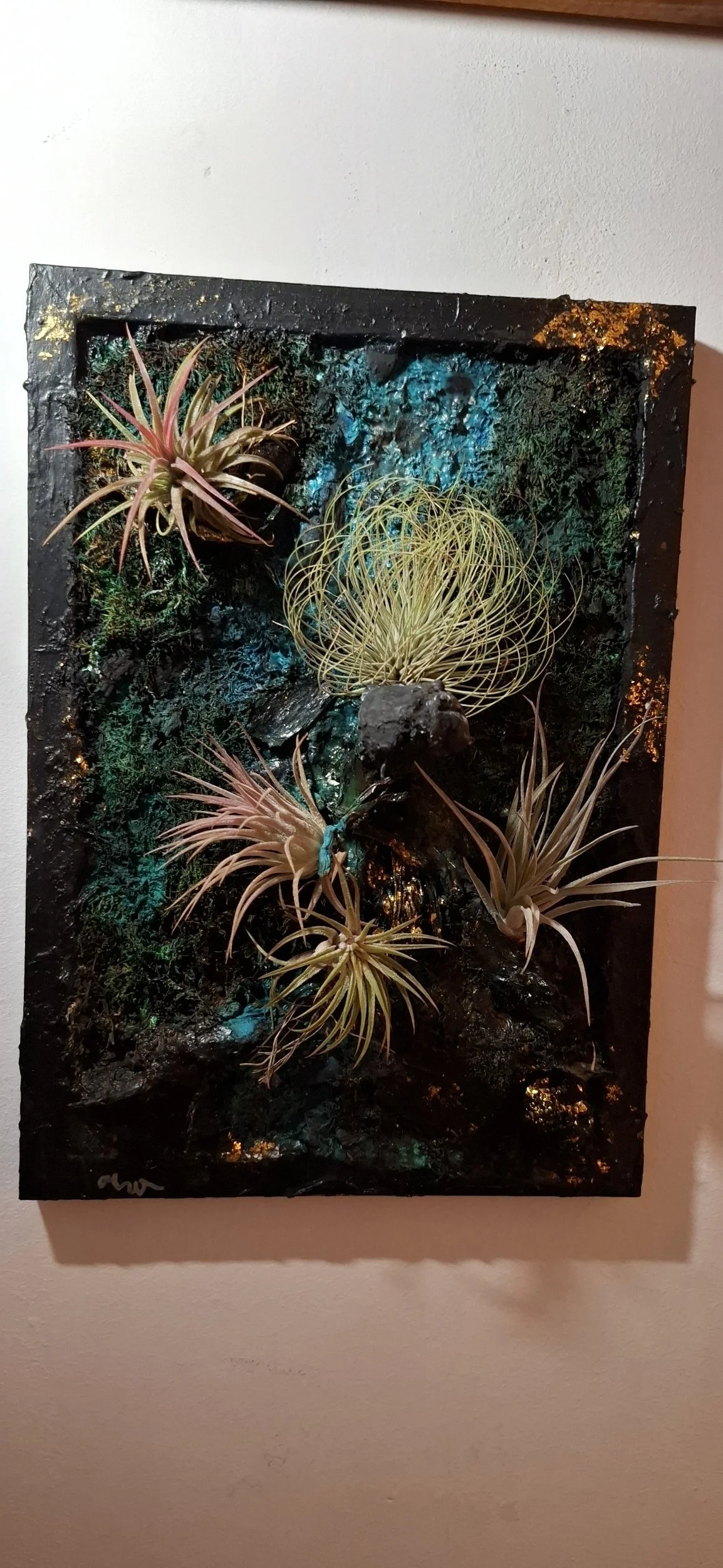 Ein Kunstwerk mit senkrecht angeordneten Sukkulenten und Flechtwerk aus Draht oder anderen Materialien, vor einem dunklen Hintergrund mit blauen und grünen Akzenten, an der Wand montiert.