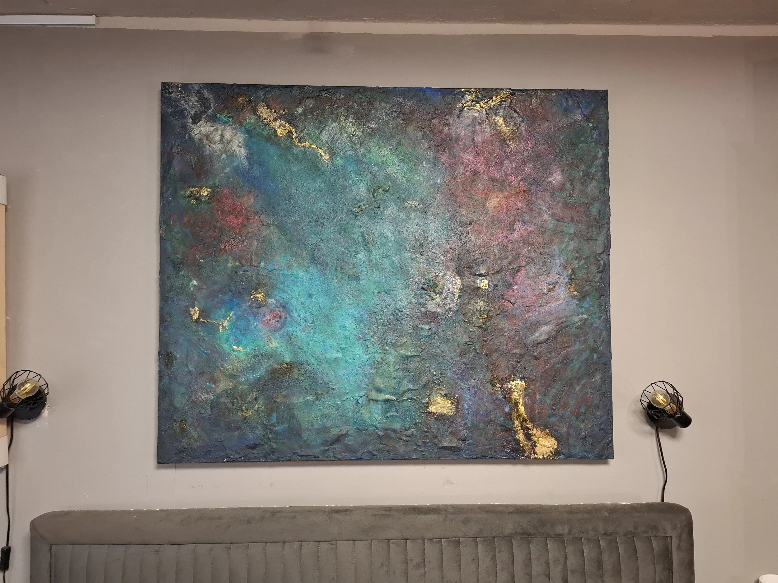 Abstraktes Wandbild mit goldenen Akzenten, blauen, grünen und rosa Farben, in einem Raum mit Wandlampen an den Seiten.