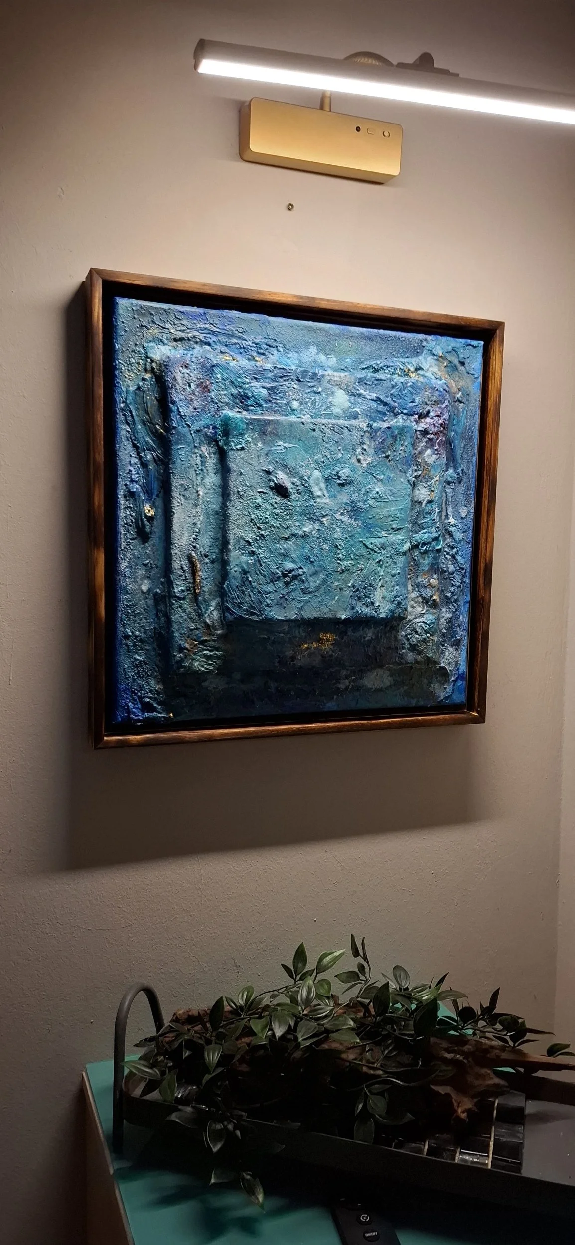 Abstraktes, texturesreiches bläuliches Gemälde in einem goldfarbenen Rahmen an einer Wand mit Deckenlicht. Darunter befindet sich eine Pflanze in einer schwarzen Kiste auf einem grünen Tisch.