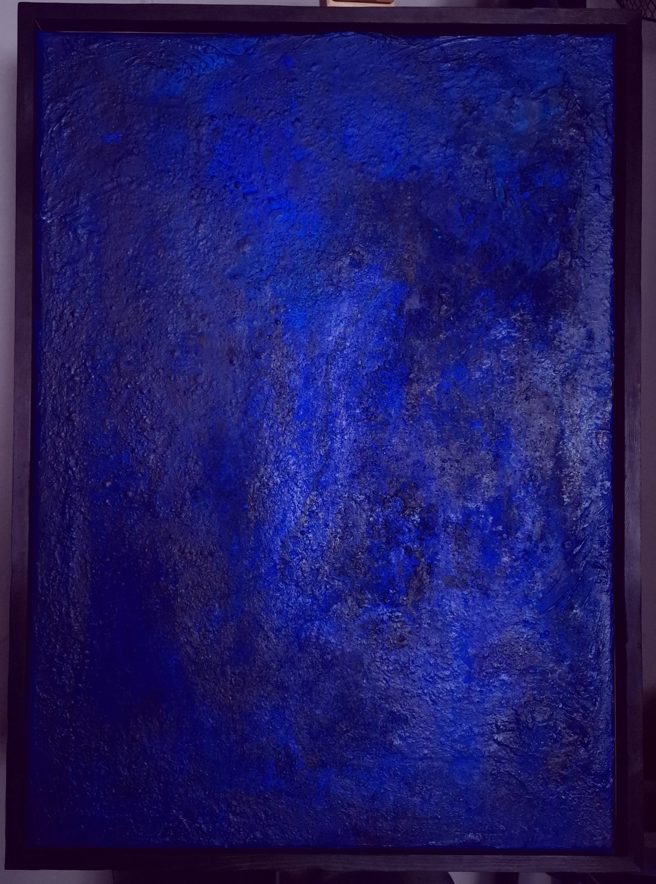 Ein großes, quadratisches Kunstwerk mit einer blauen, texturierten Oberfläche, eingerahmt in einen dunklen Rahmen, abgelichtet in einer Galerie.