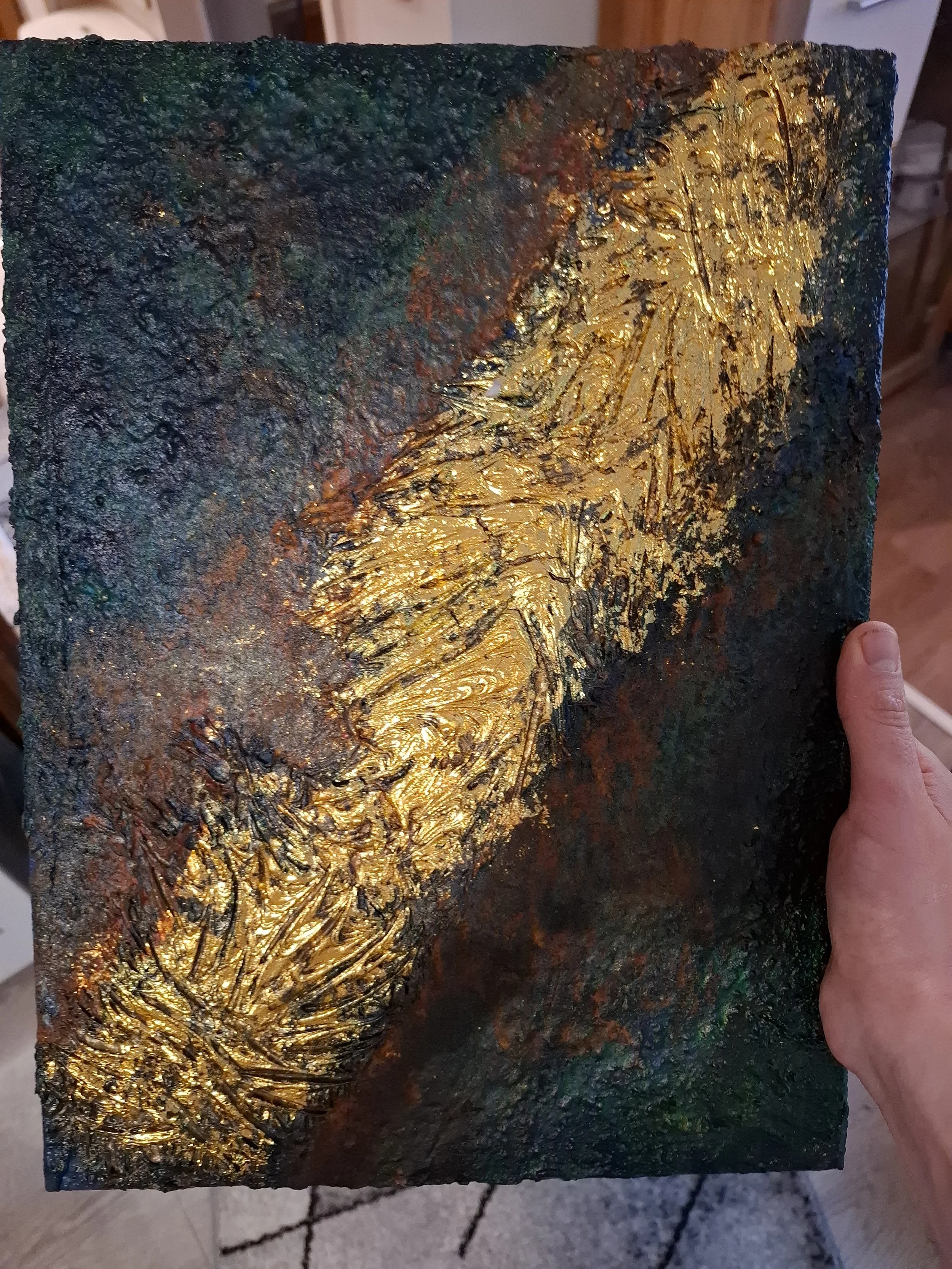 Ein handgefertigtes Kunstwerk mit einer schwarzen, grüngrauen Grundierung und goldenen, strukturierten Akzenten in abstraktem Design.