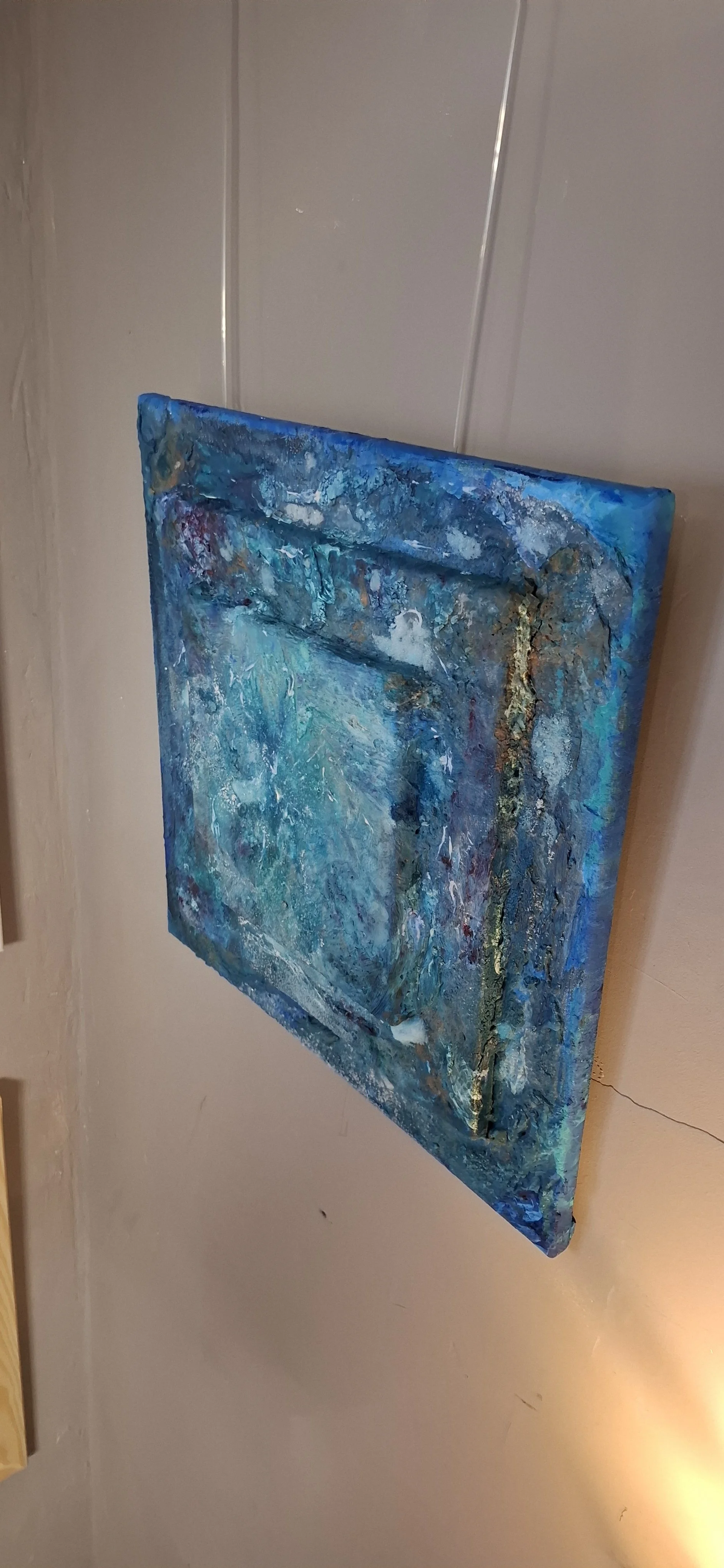 Abstraktes Gemälde mit verschiedenen Blautönen und goldenen Akzenten, aufgehängt an einer Wand.