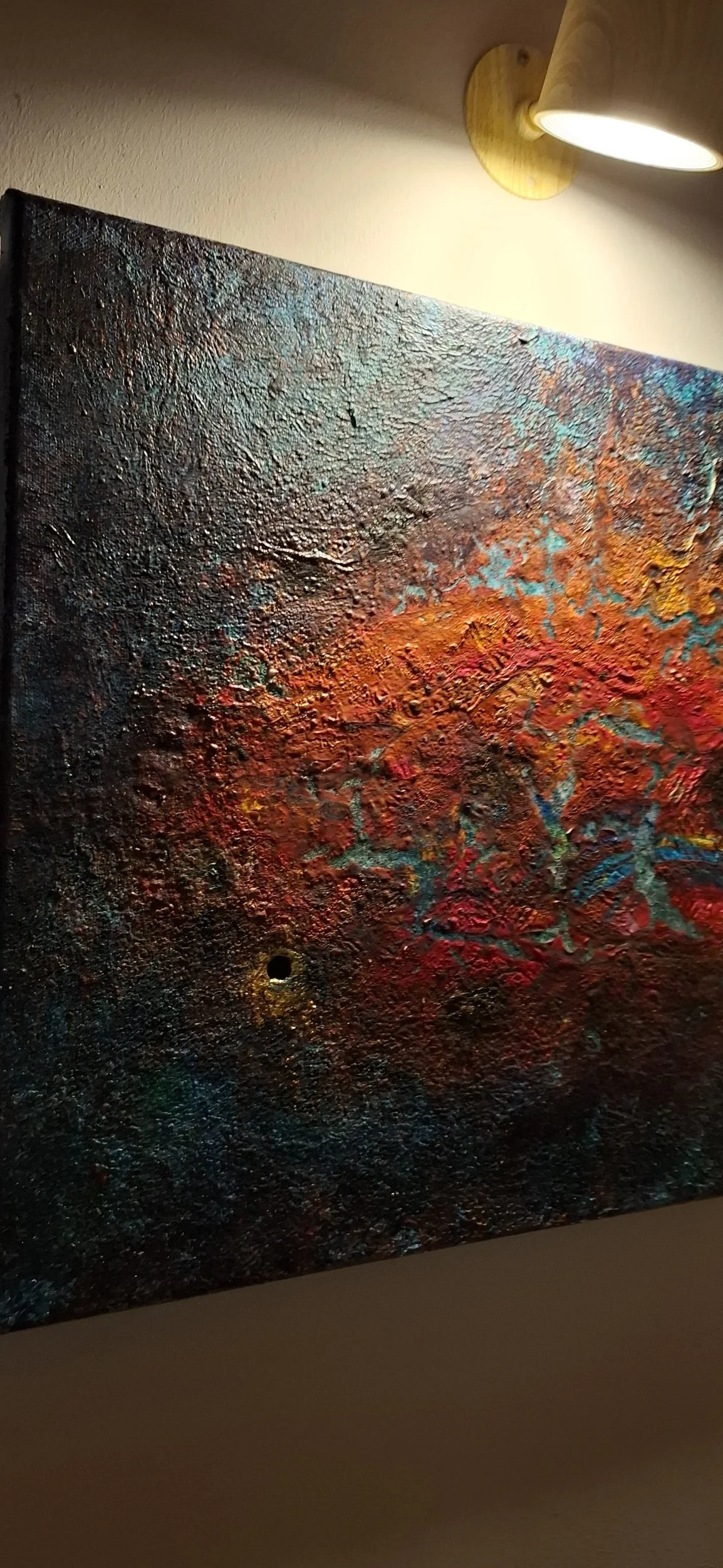 Ein abstraktes, farbenfrohes Gemälde mit einer Textur, das an einen Sonnenuntergang oder eine Feuerlandschaft erinnert, beleuchtet von einer Wandlampe.