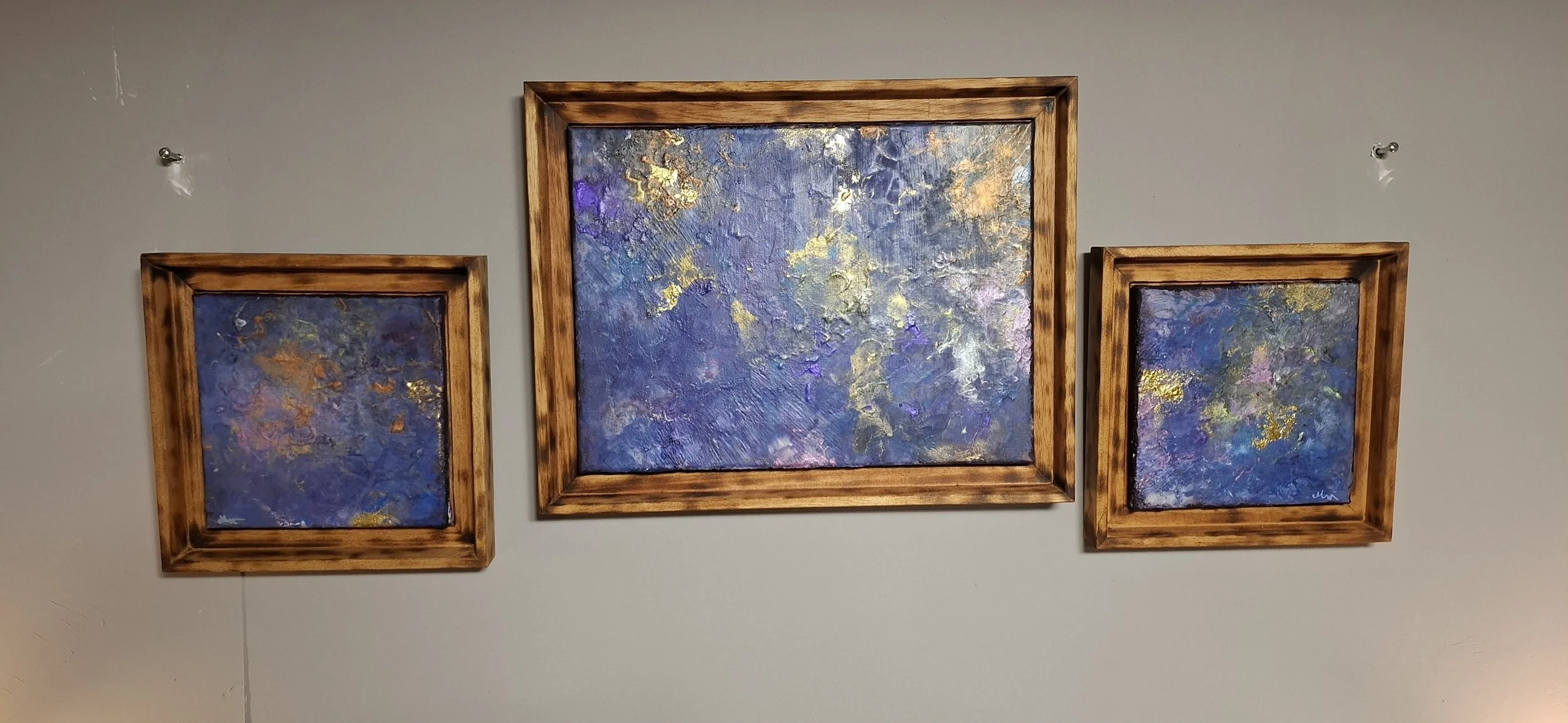 Drei bunte, abstrakte Gemälde in Holzrahmen an einer grauen Wand.