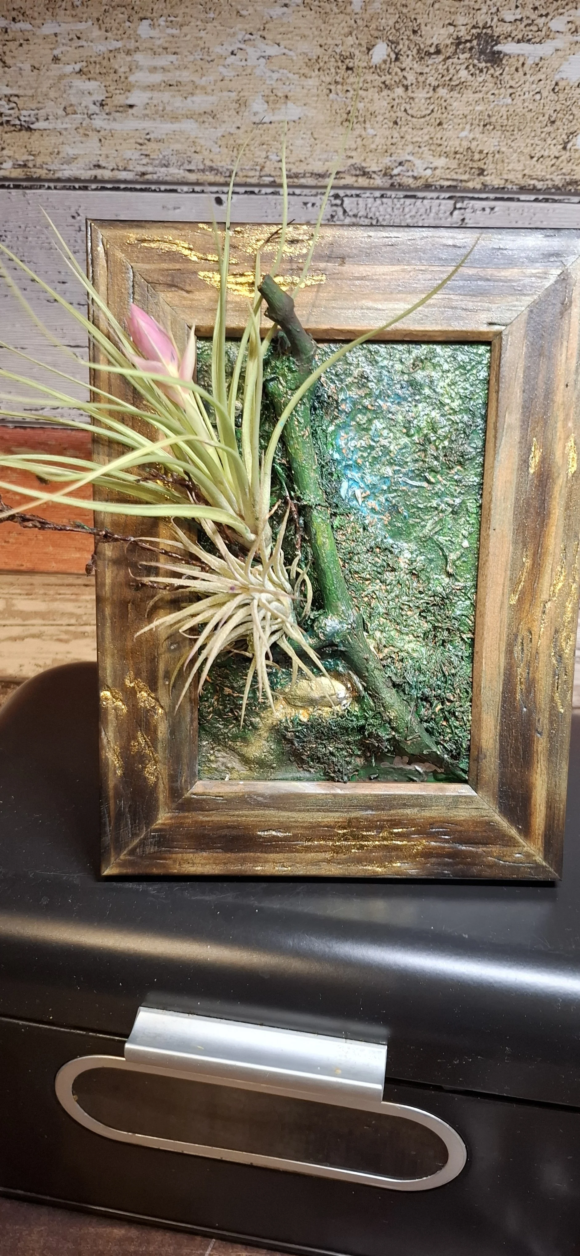 Ein Holzrahmen mit einer 3D-Ikone aus Moos, Holz und Pflanzen in Pastellfarben.