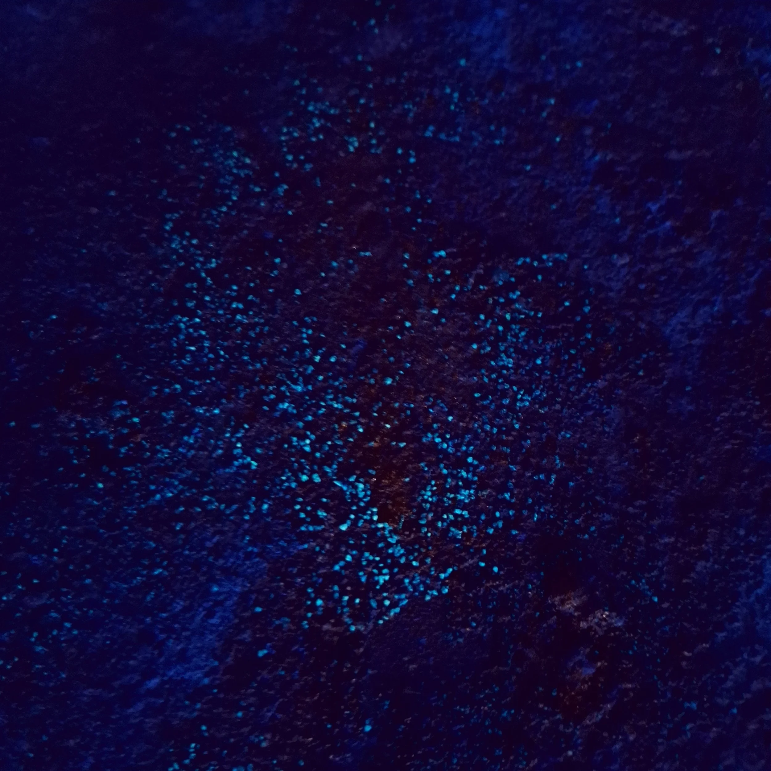 Nahaufnahme von bläulichen Glitzerelementen oder -pigmenten auf einer dunklen Oberfläche, die im Dunkeln leuchten.