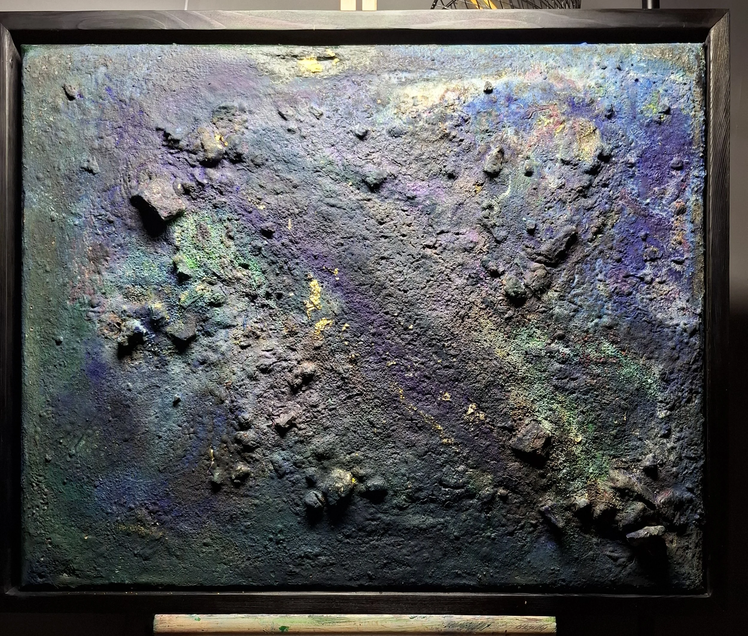 Eine große, quadratische Kunstinstallation mit einer Oberfläche aus gekrümmter, rauer Textur in dunklen, metallischen Farben. Umgeben von einem schwarzen Rahmen, auf dem oberen Rand befindet sich eine dunkle Holzleiste.