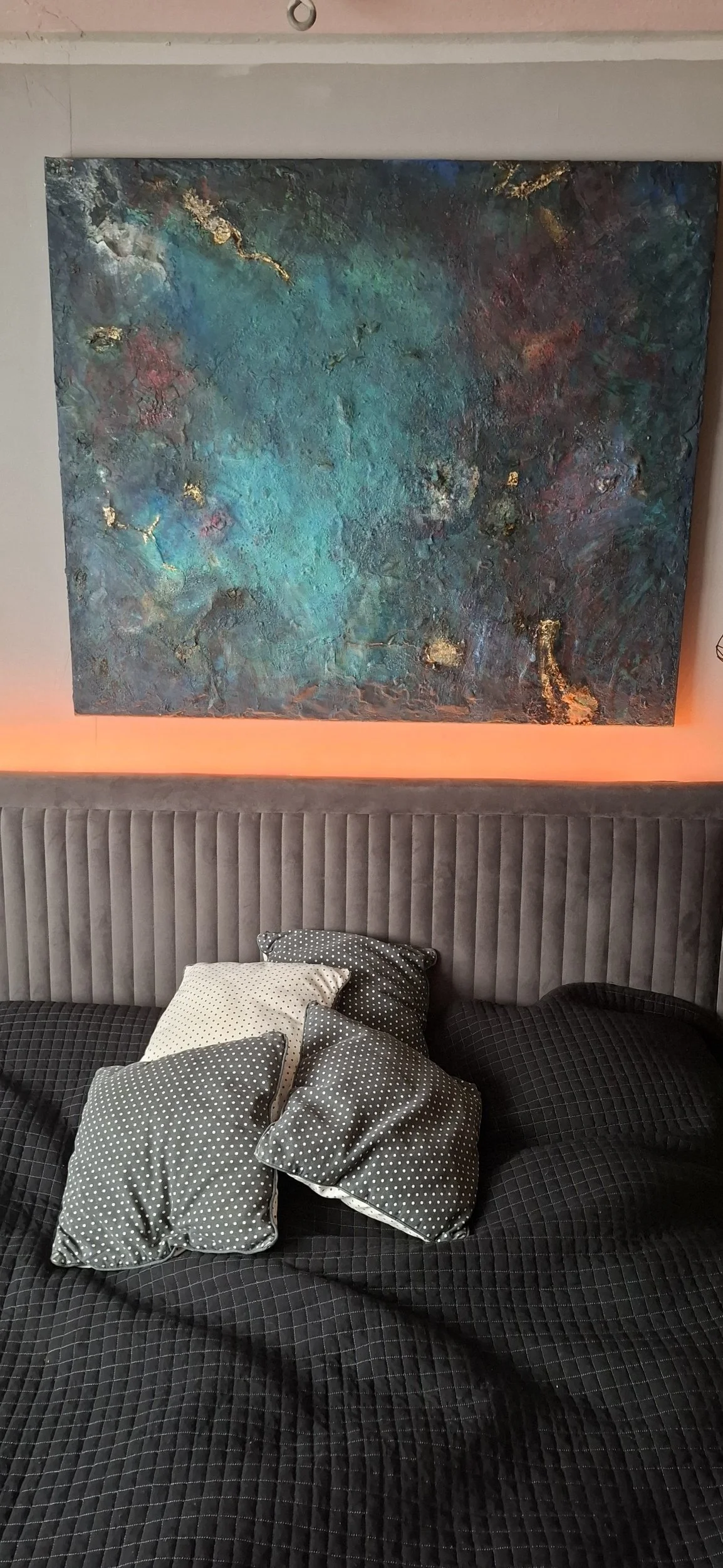 Ein modernes Schlafzimmer mit einem grauen, gesteppten Kopfteil, schwarzen Bettdecken und mehreren Mustertüchern in Grau und Weiß. Über dem Bett hängt ein großes, abstraktes Gemälde mit Blau-, Grün- und Goldtönen, das an die Wand gemalt ist.