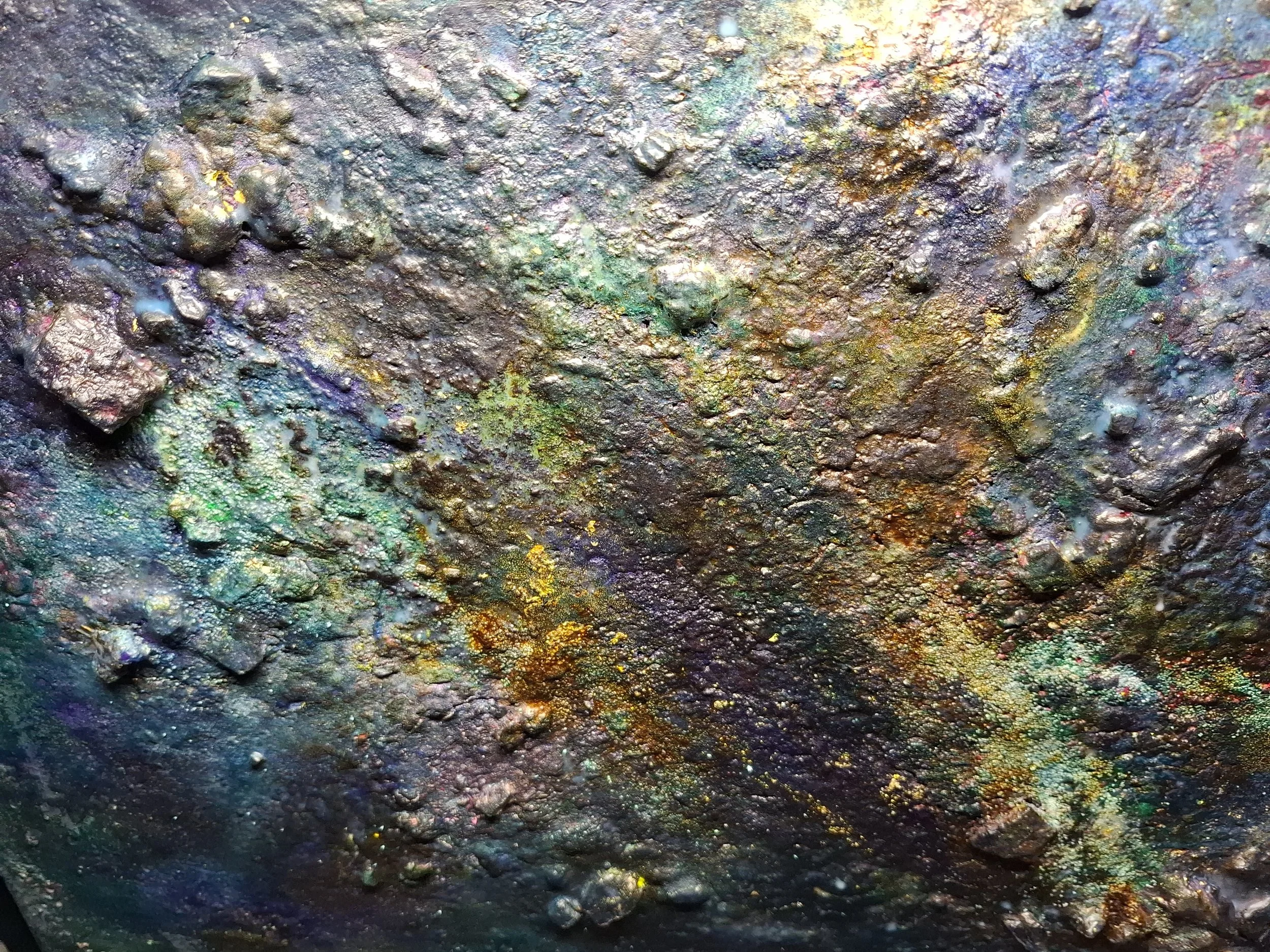 Ein farbenfrohes, metallisches, ungleichmäßiges Oberflächenmuster mit schillernden Farben wie Gold, Grün, Blau und Violett, das wie Lack oder Aquarell wirkt.