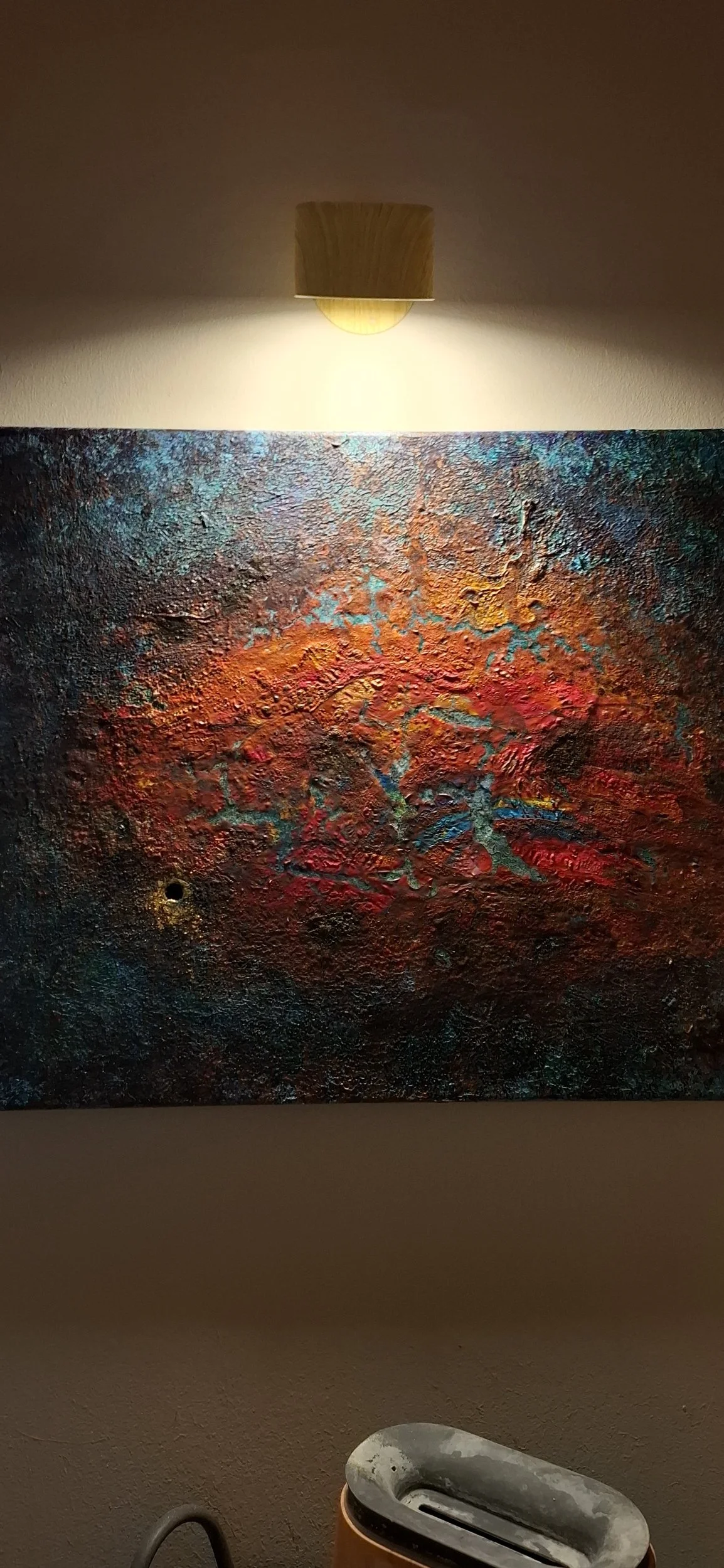 Ein abstraktes, texturiertes Gemälde mit dominanten Rottönen, blauen und orangefarbenen Akzenten, beleuchtet von einer Wandlampe, mit einem Staubsauger im Vordergrund.