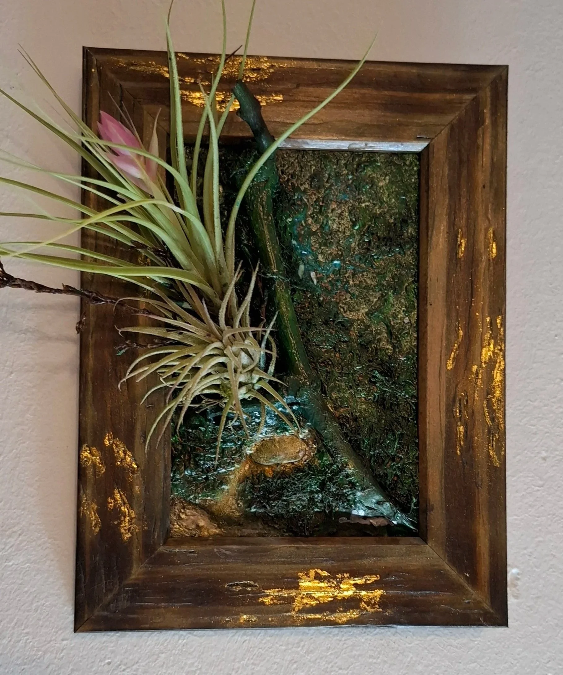 Ein Wandkunstwerk mit einer metallischen Skulptur eines Rehs und luftigen Pflanzen, eingebettet in einem Holzbilderrahmen mit goldenen Akzenten.