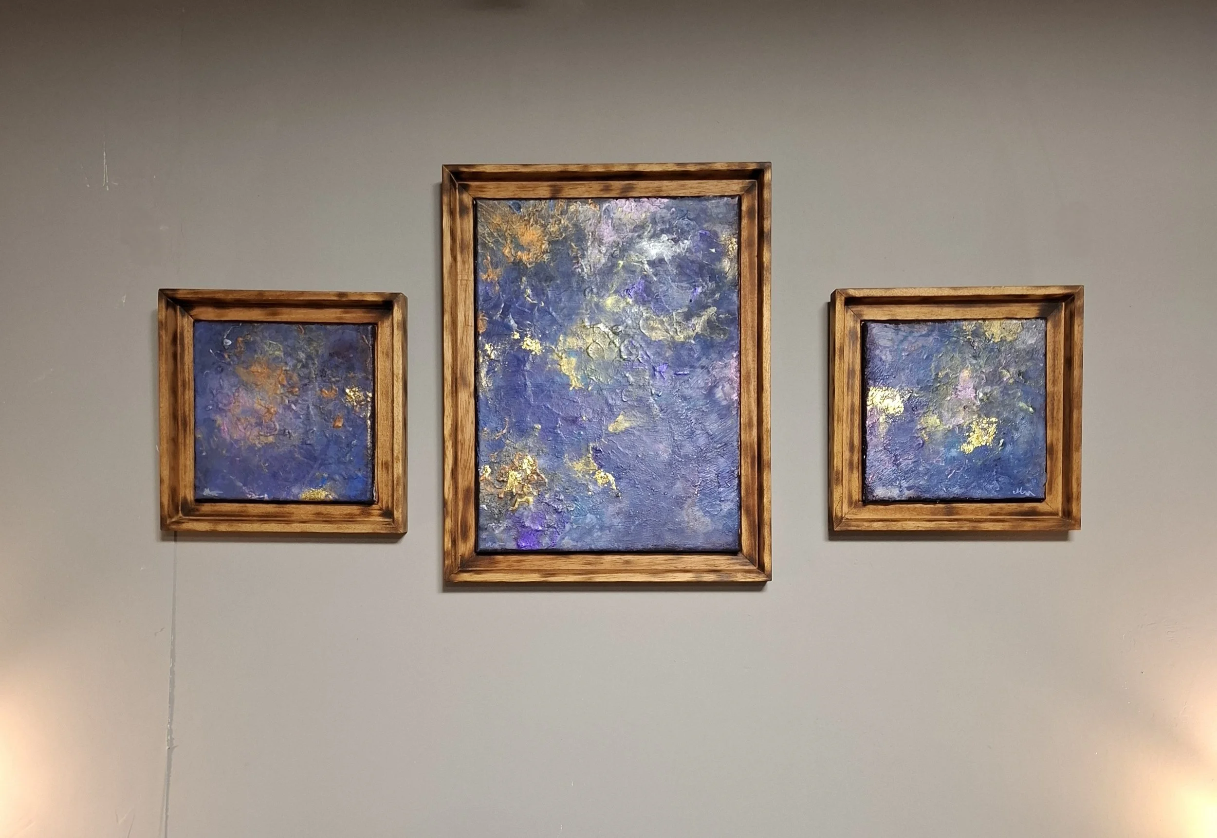 Drei abstrakte Gemälde mit blauen und goldenen Akzenten in Holzrahmen an einer grauen Wand.