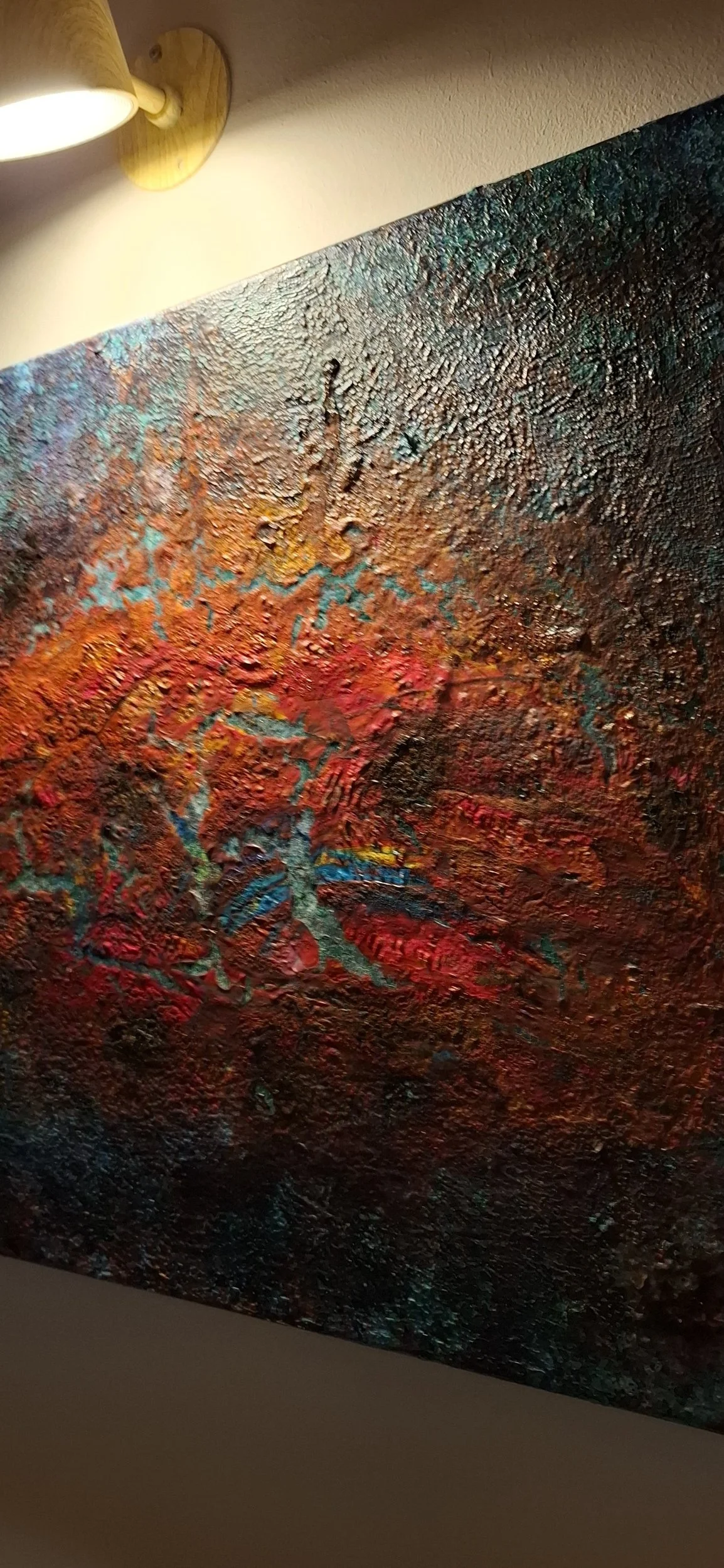 Ein abstraktes, farbenfrohes Gemälde mit strukturierter Oberfläche und lebhaften Rot-, Blau-, Orange- und Gelbtönen, beleuchtet von einer Wandlampe.