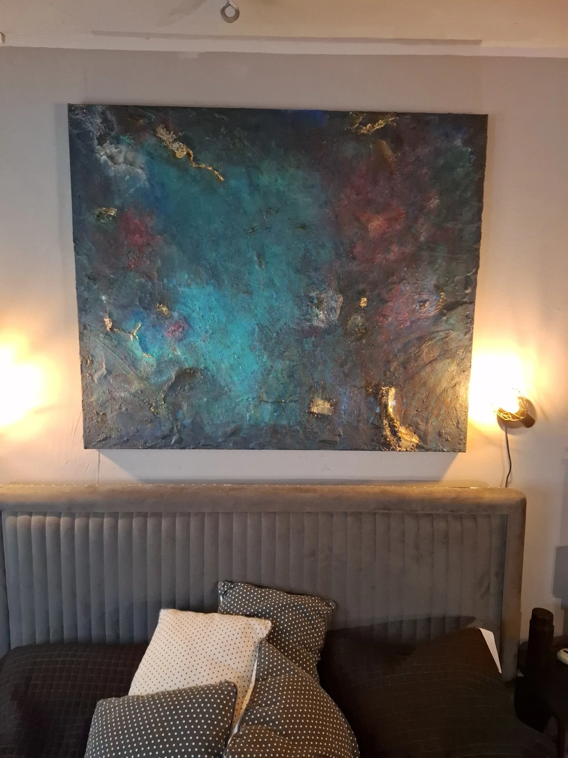 Ein Wohnzimmer mit einer gepolsterten Sitzbank, mehreren Polsterkissen mit Punkten, einem gemalten Bild an der Wand und einer Stehlampe, die das Bild beleuchtet.