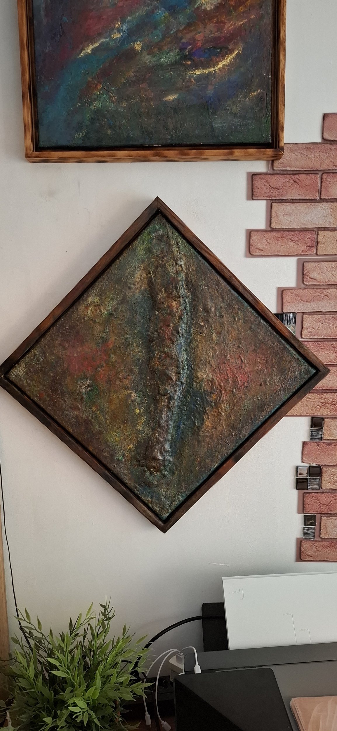 Zwei abstrakte Gemälde an einer weißen Wand, eines in einer quadratischen Holzrahmen, das andere in einem diagonalen Holzrahmen, neben einer roten Backsteinwand und einem Schreibtisch mit Pflanzen und elektronischen Geräten.
