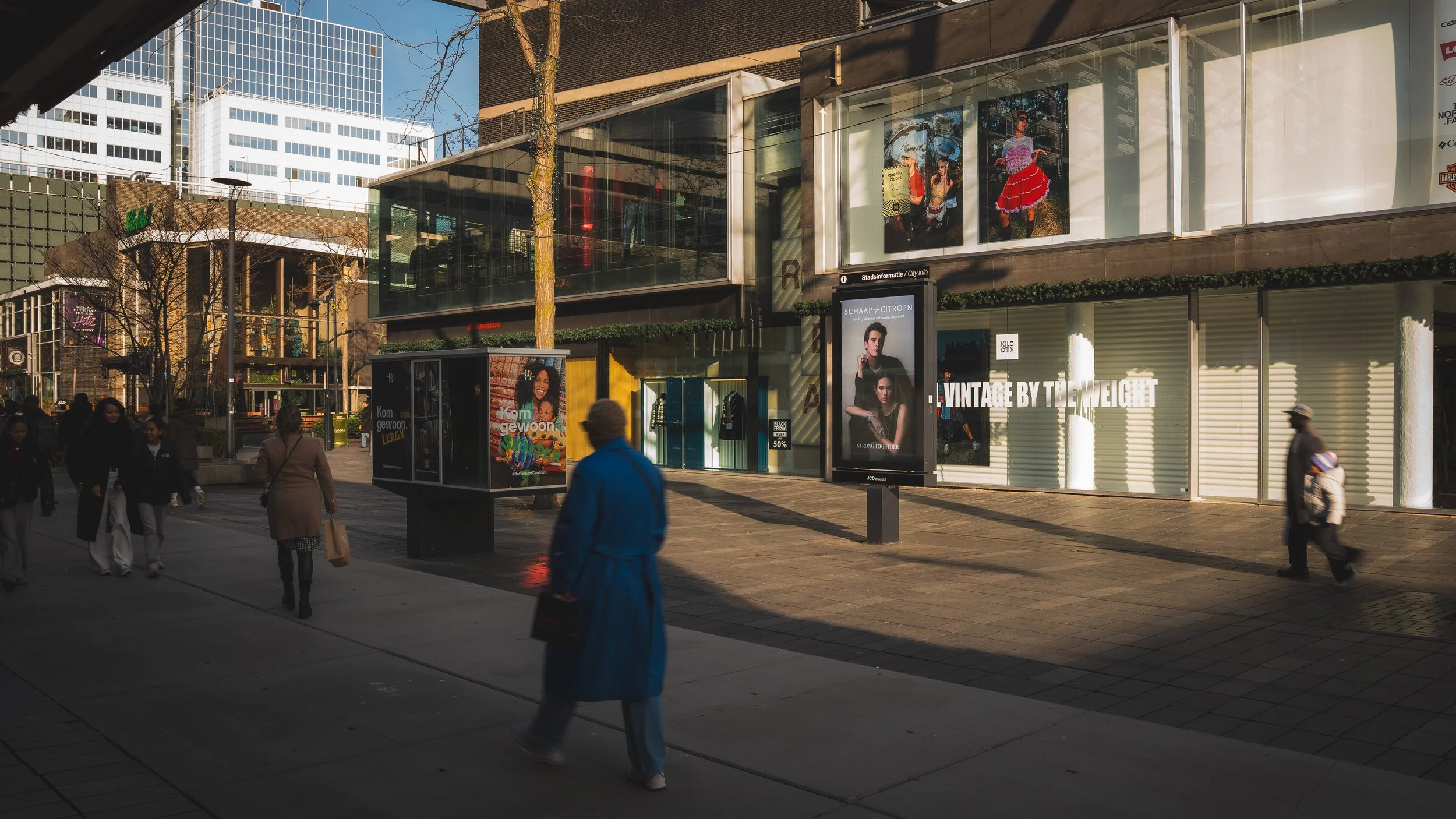 Straatbeeld met mensen die wandelen, moderne winkelgevels met posters en digitale advertenties, en hoge gebouwen op de achtergrond.