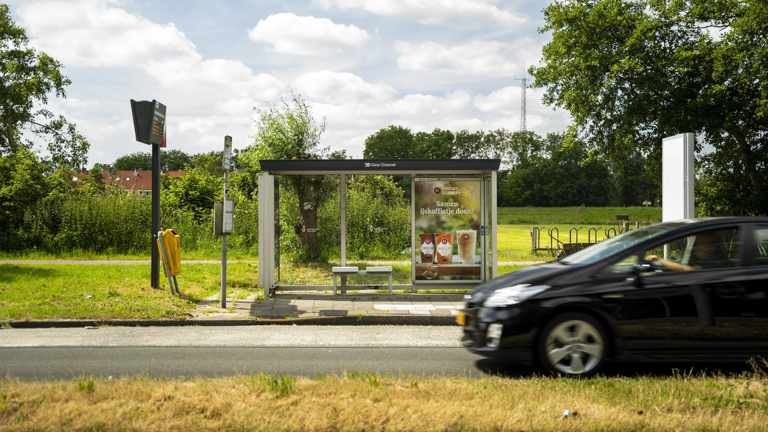 Bushalte met een reclamebord en een bankje, naast de straat met een zwarte auto die passeert, omgeven door groene bomen en gras.
