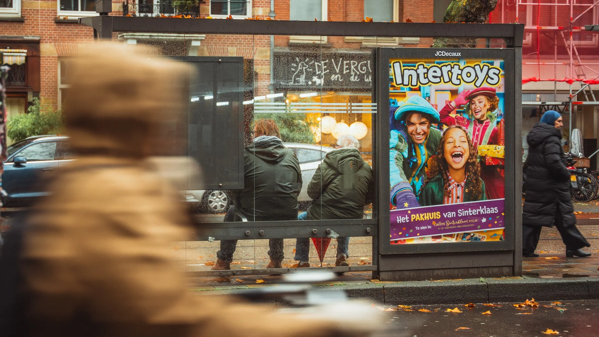 Drie mensen wachten bij een bushokje op een regenachtige dag. Op de muur achter hen hangt een poster voor het speelgoedmerk Intertoys en een reclame voor het Pakhuis van Sinterklaas.