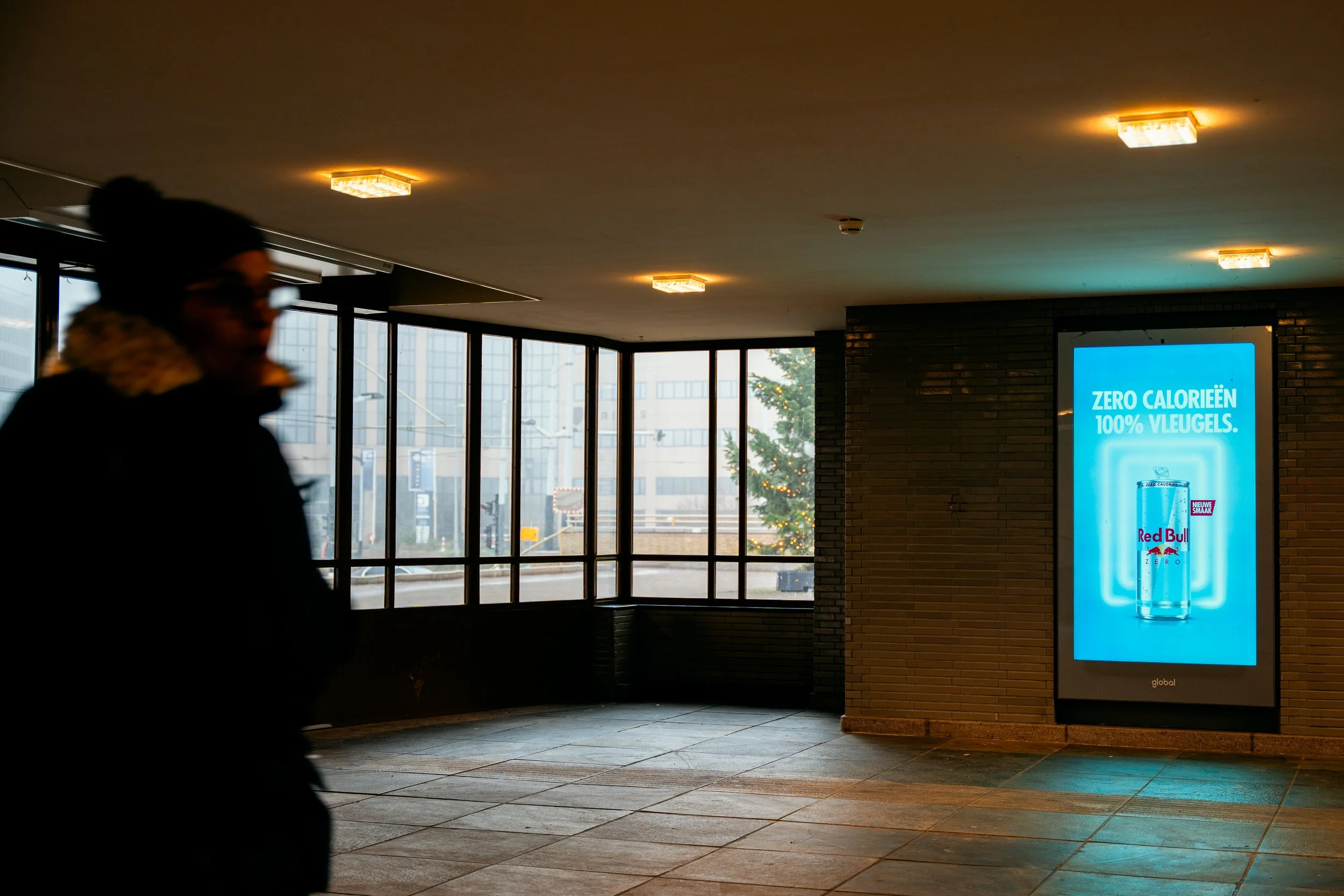 Een silhouet van een persoon die langs een digitale reclamezuil loopt binnen in een gebouw.