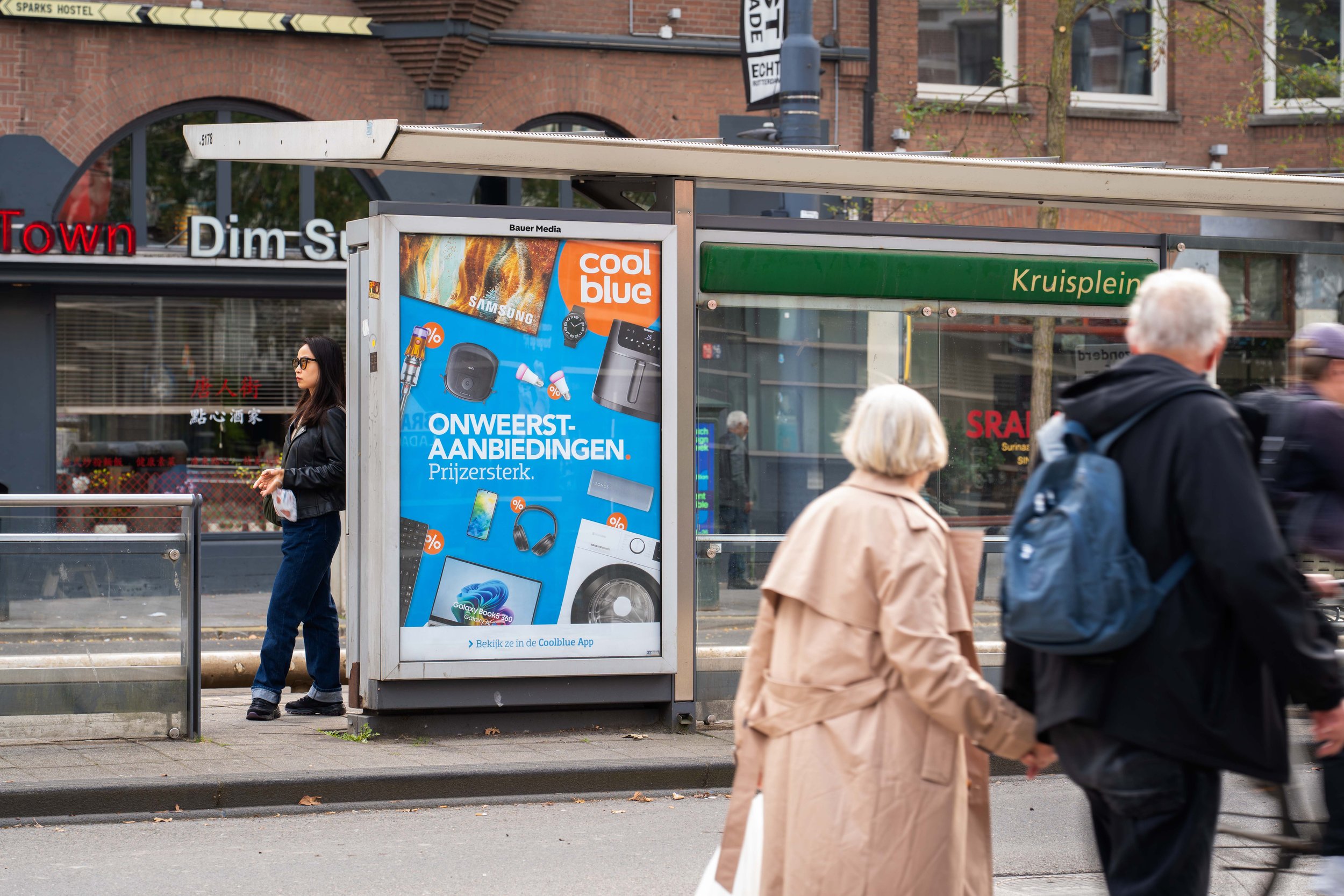 Mensen wachten bij een bushalte met een reclamebord en een doorzichtige overkapping. De achtergrond toont een gebouw met een boog en ramen, en een groene winkelzeil met de naam 'Kruisplein'.