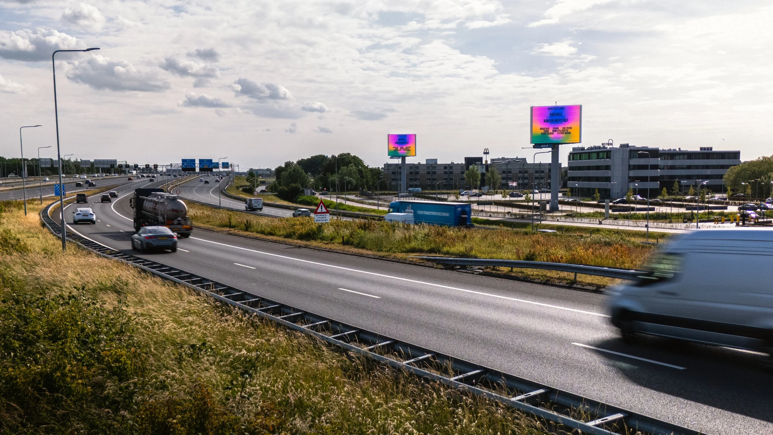 Snelweg met rijdende voertuigen en elektronische billboards op achtergrond onder een bewolkte hemel.