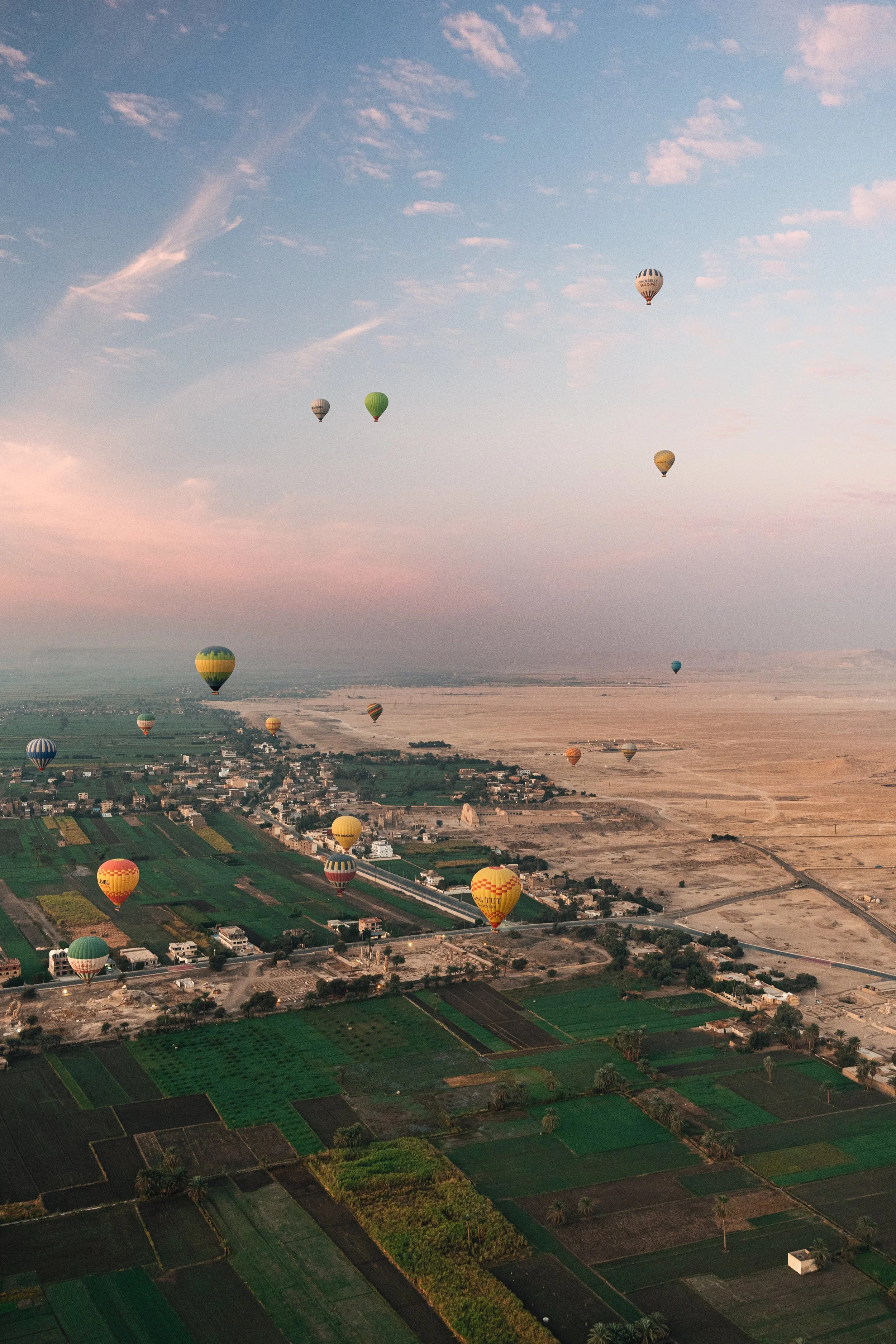 Verschillende kleurrijke helikopters zweven boven een uitgestrekt landschap met velden, huizen en een rivier, onder een blauwe lucht met verspreide wolken.