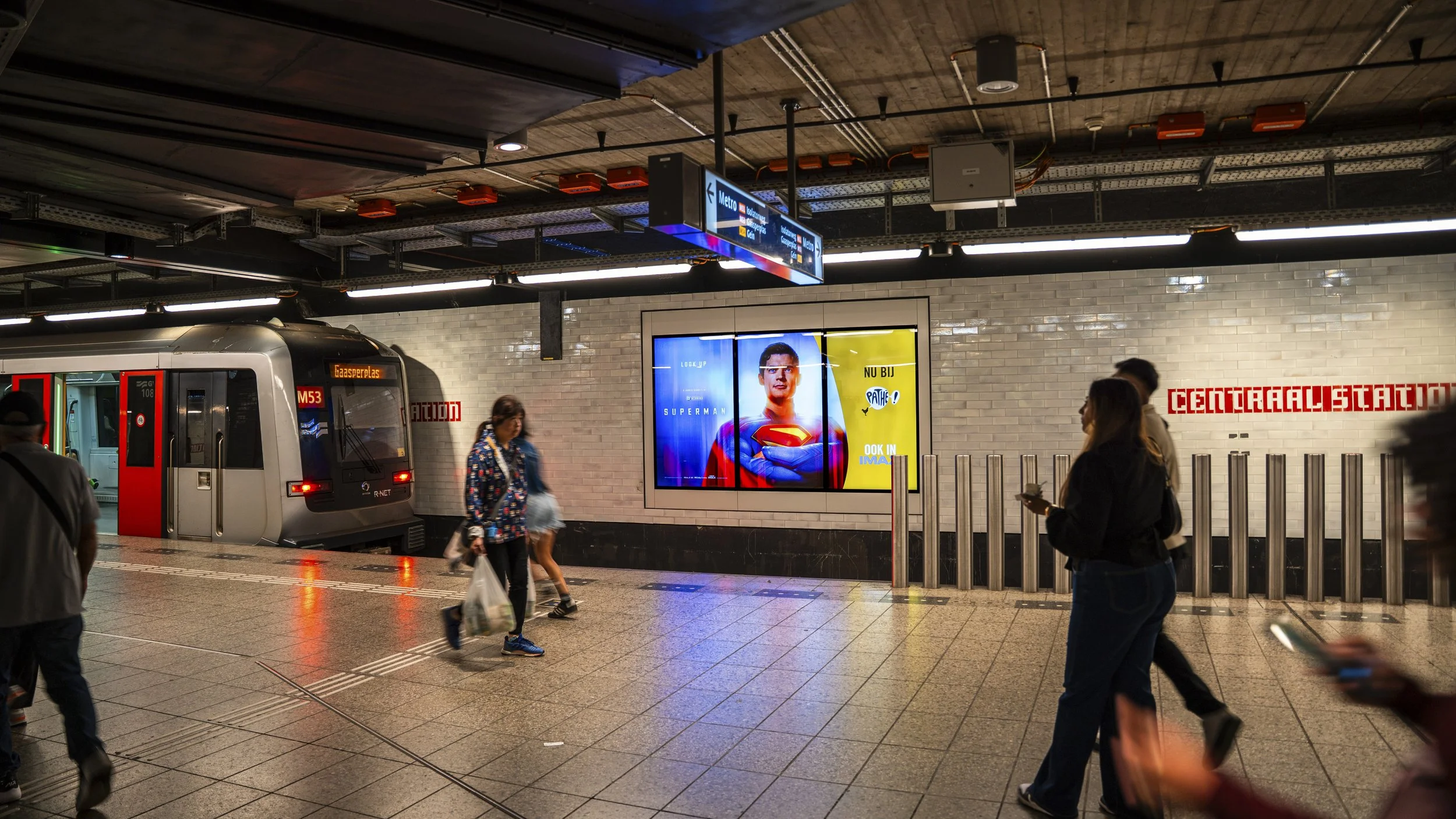 Een metrostation met mensen die wachten, een metro die binnenkomt en een digitale reclamedisplay met Superman erop.