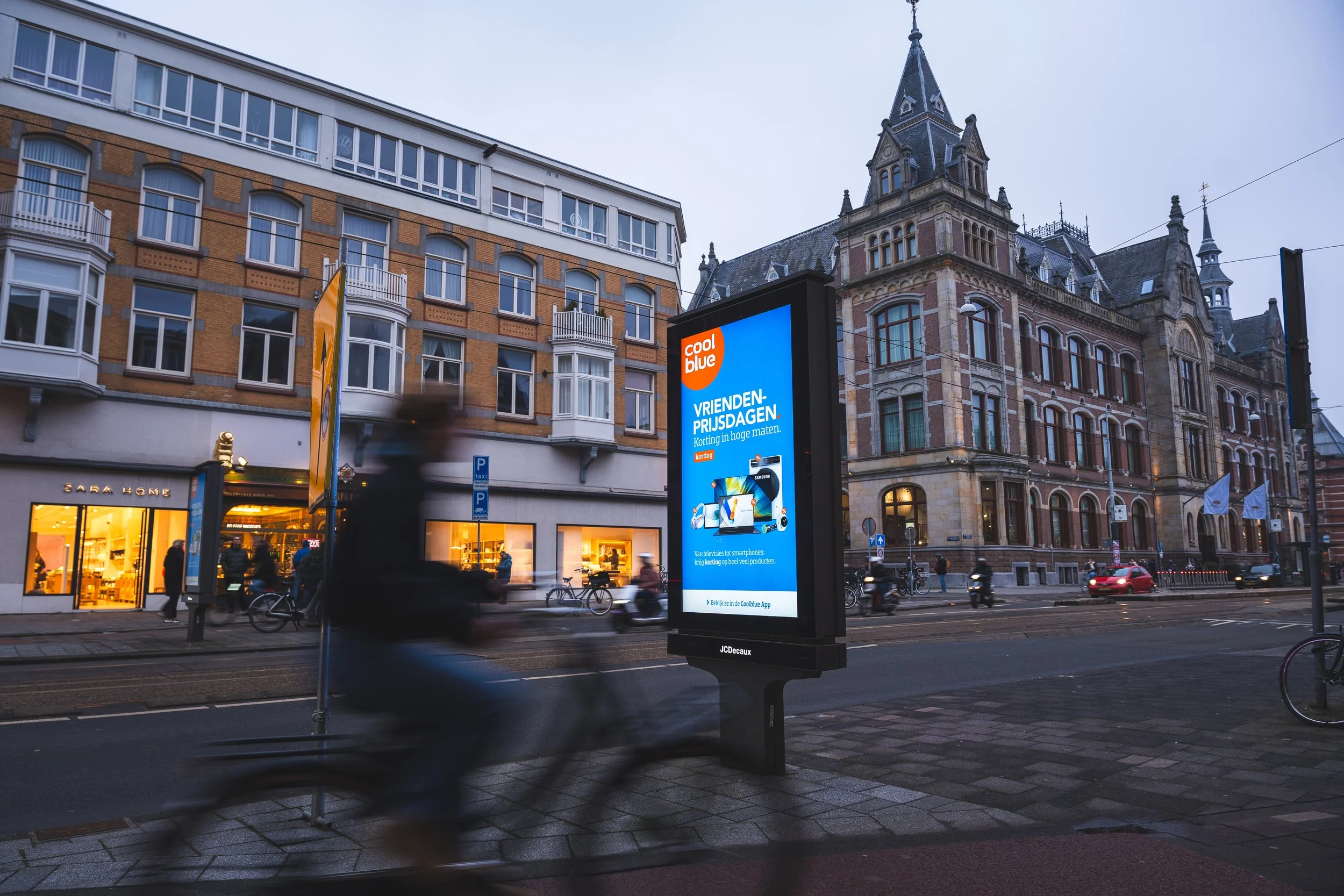 Schermafbeelding van een straat in de avond met een displayadvertentie voor Coolblue. Mensen fietsen en lopen langs de straat, met een historisch gebouw op de achtergrond.