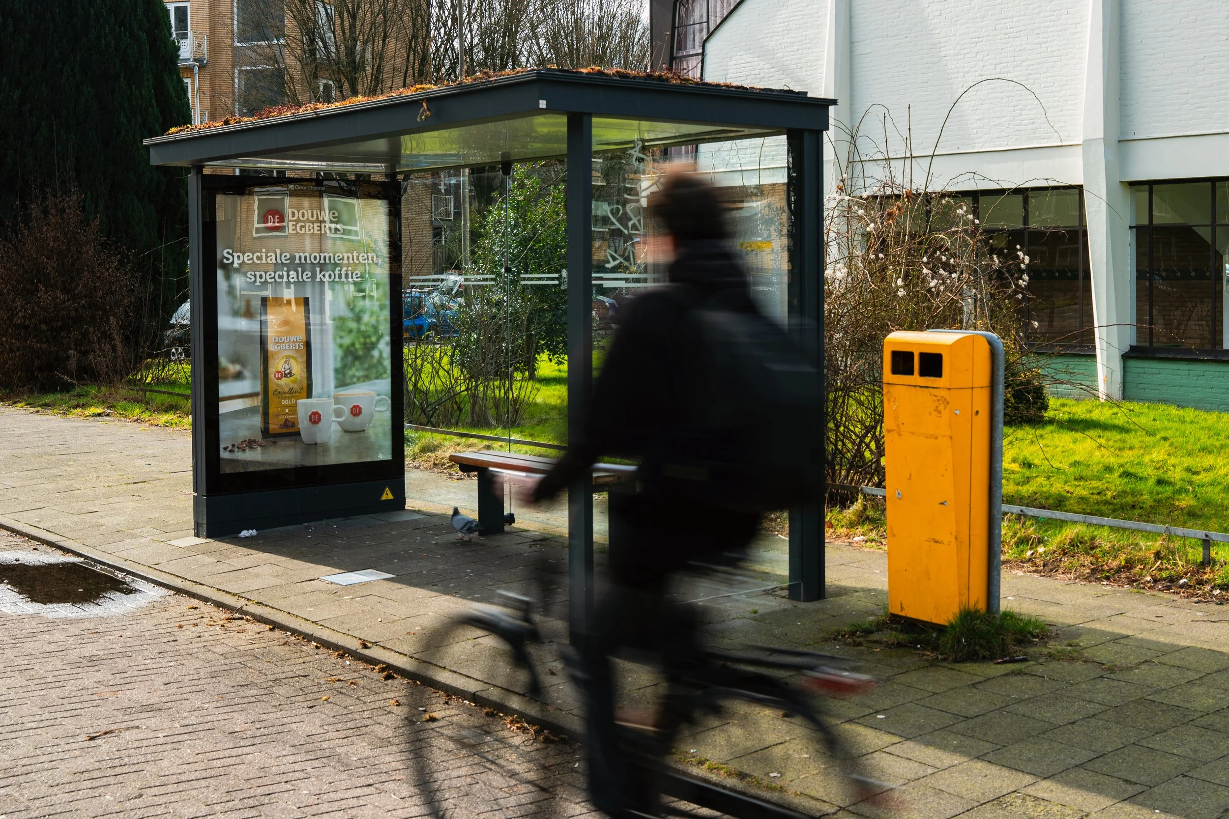 Een persoon fietst langs een bushokje met een advertentie dat reclame maakt voor Douwe Egberts koffie, op een straat met een geel prullenbak en een groene tuin op de achtergrond.