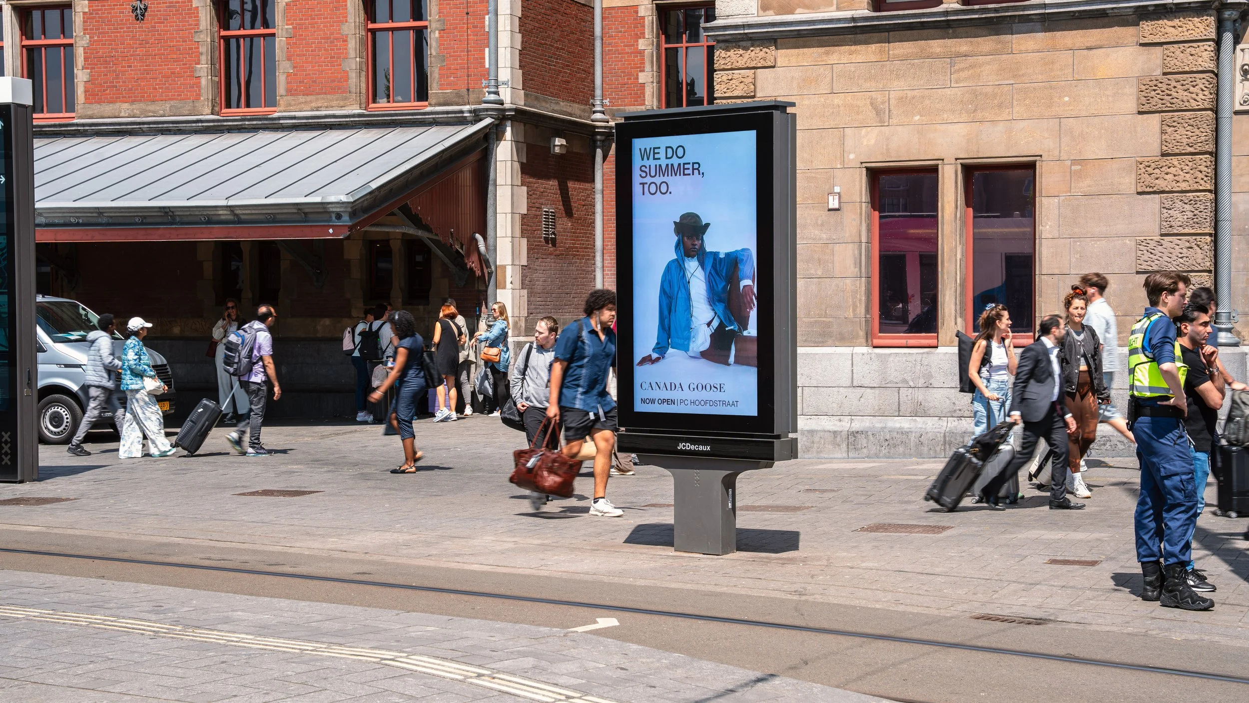 Een drukke straat met mensen die lopen met koffers en tassen. Op de achtergrond een gebouw met roodbruine bakstenen en grote ramen. In het midden een digitale billboard met een advertentie voor Canada Goose dat zegt: 'We do summer, too.'