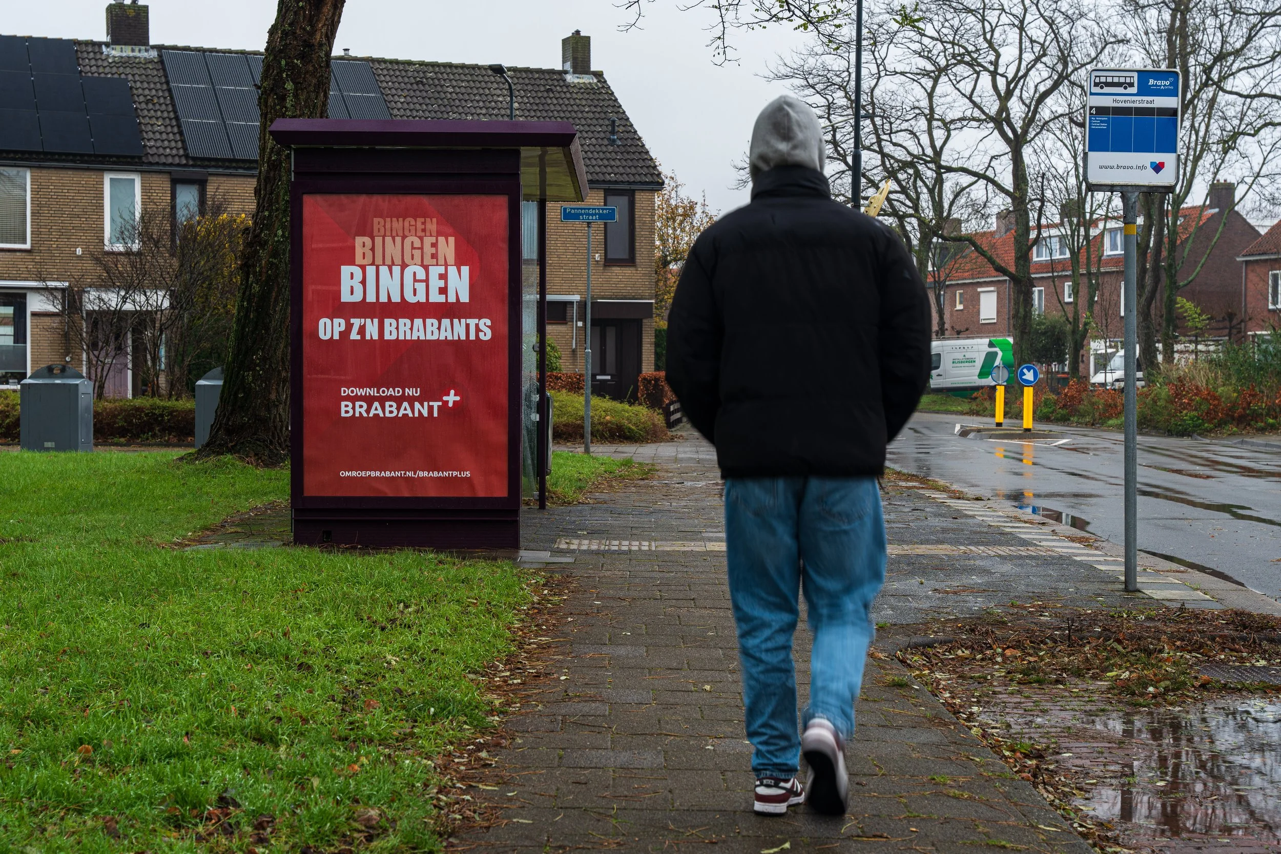 Een persoon in een zwarte jas en lichte spijkerbroek loopt op een regenachtige stoep in een woonwijk, met een waarschuwingsbord en een reclamebord voor Brabant op de achtergrond.