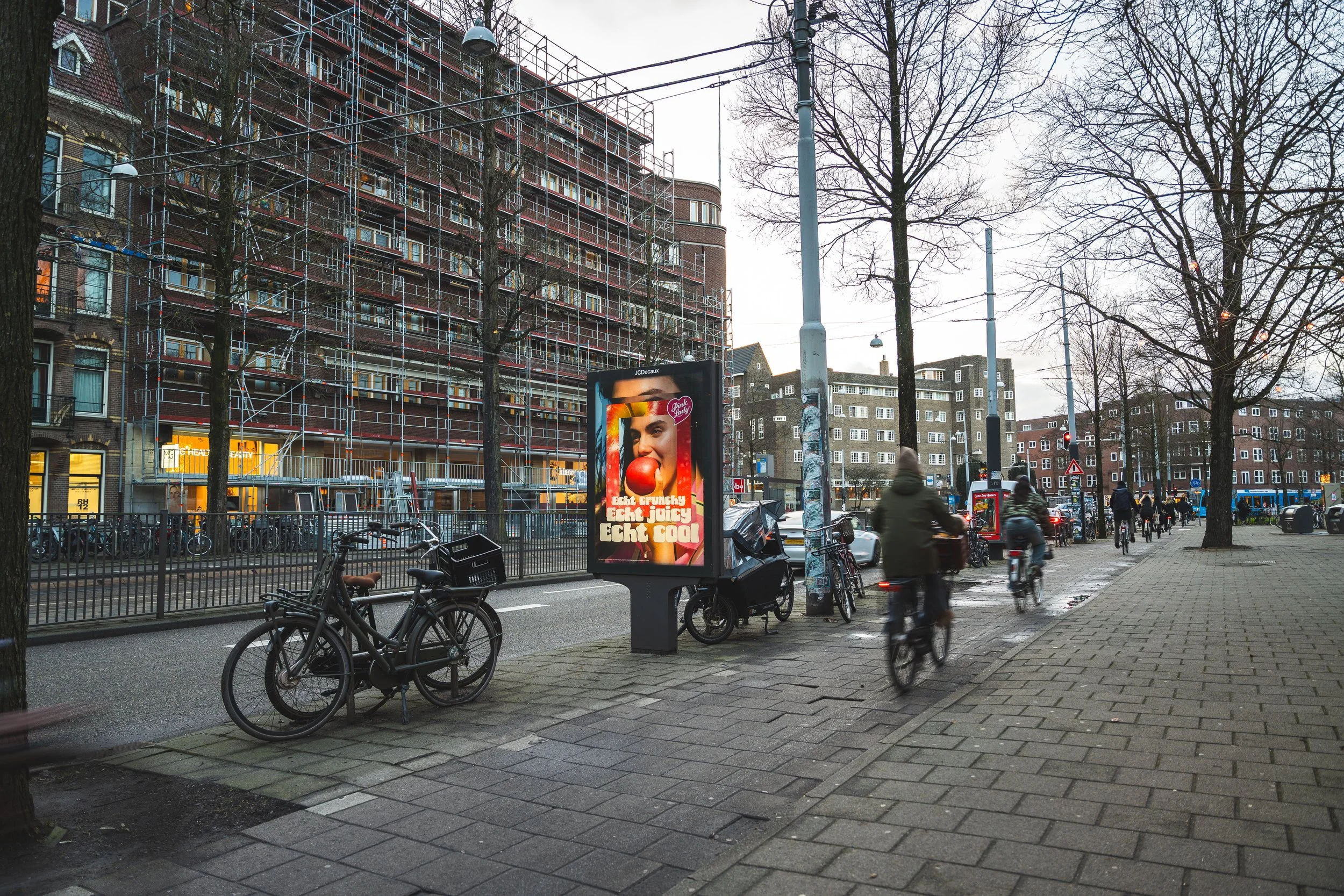 Straat met fietsen, mensen op de fiets, een advertentiebord met een vrouw en tekst, bomen, gebouwen en een verkeerslicht