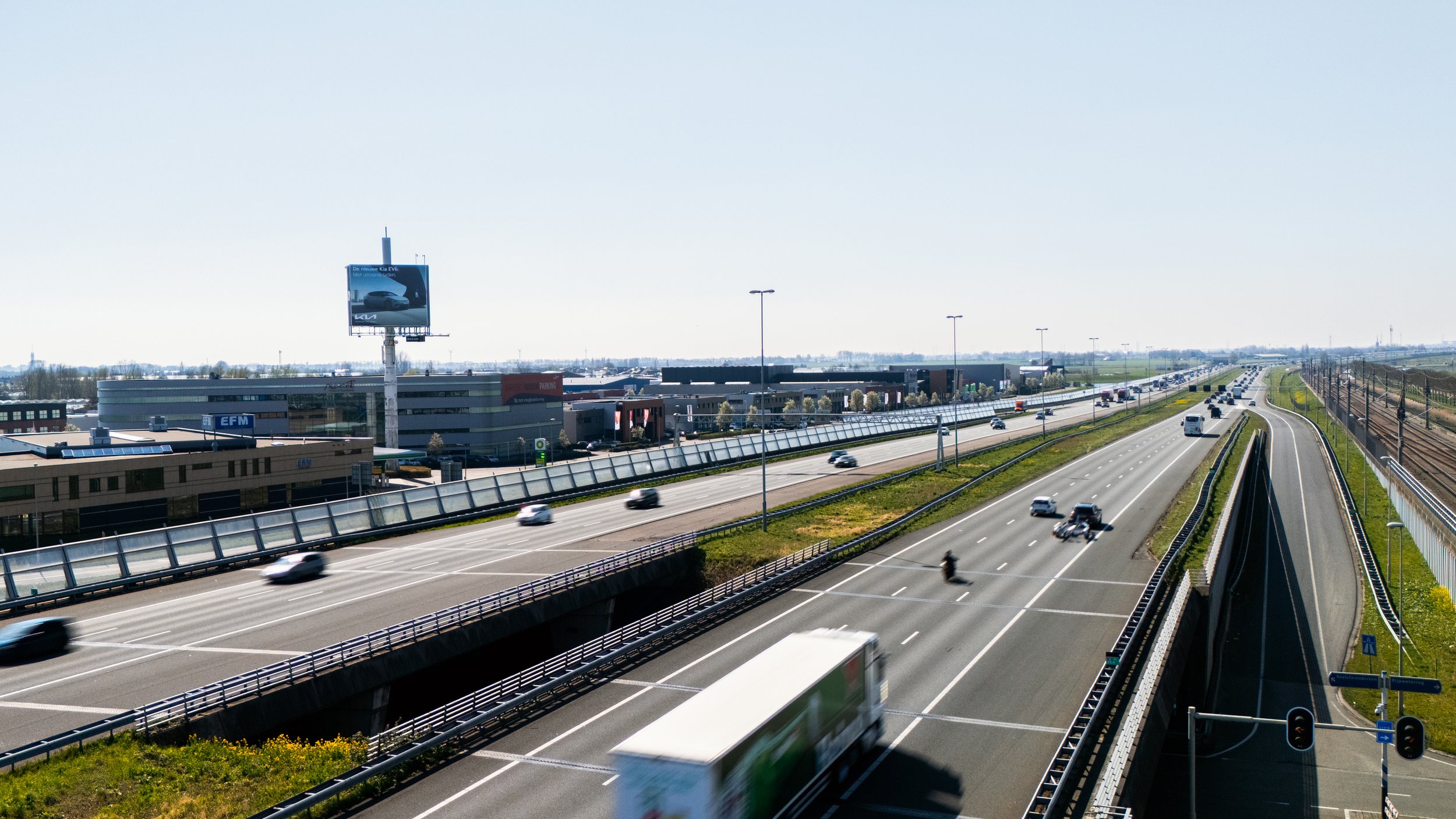 Snelweg met meerdere rijstroken vol auto's en vrachtwagens, met een bushalte aan de rechterkant en een winkelcentrum op de achtergrond, onder een heldere blauwe hemel.