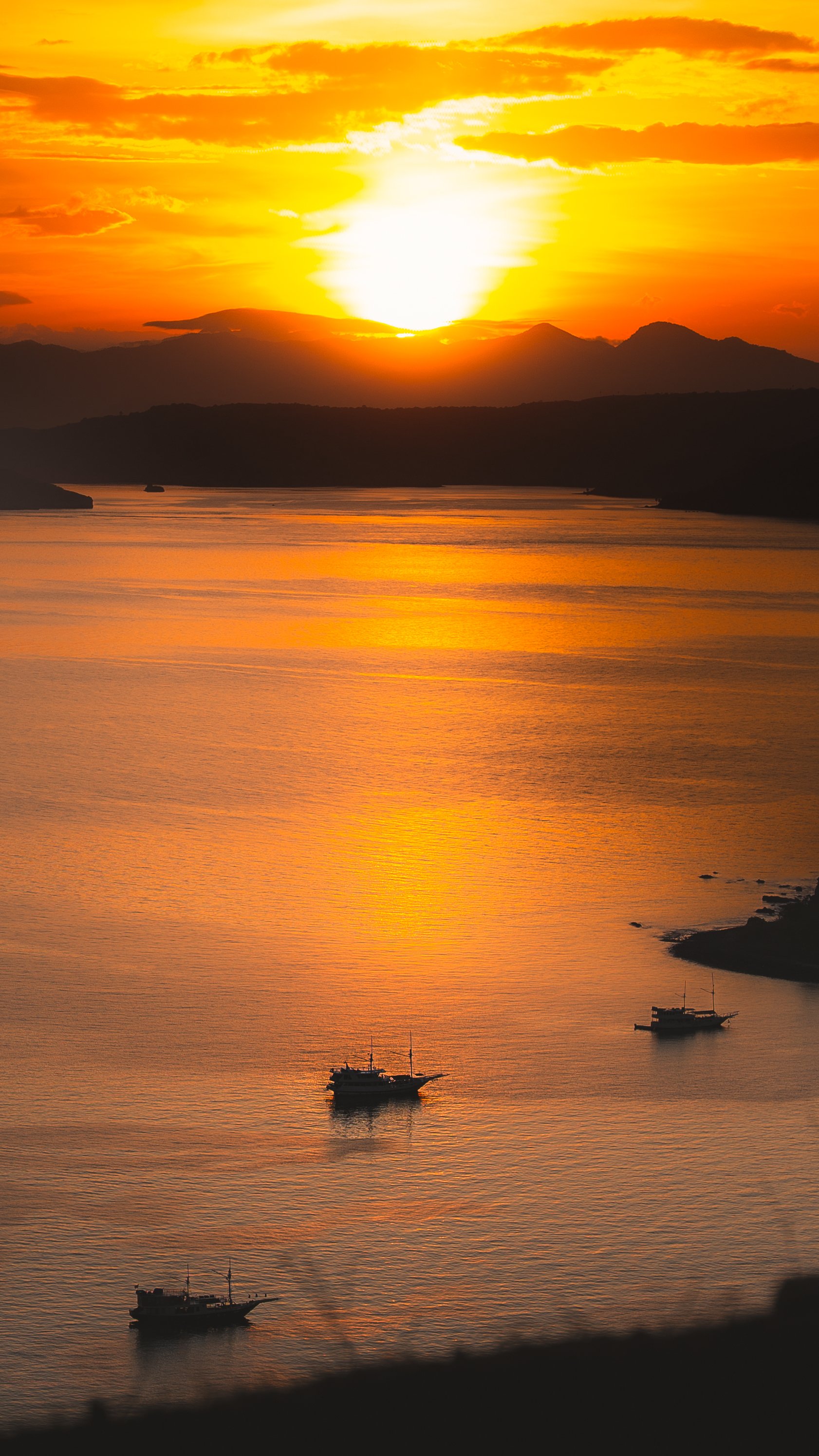 Een zonsondergang boven een kalm meer met vier boten en bergen op de achtergrond, alles getint in oranje en goud