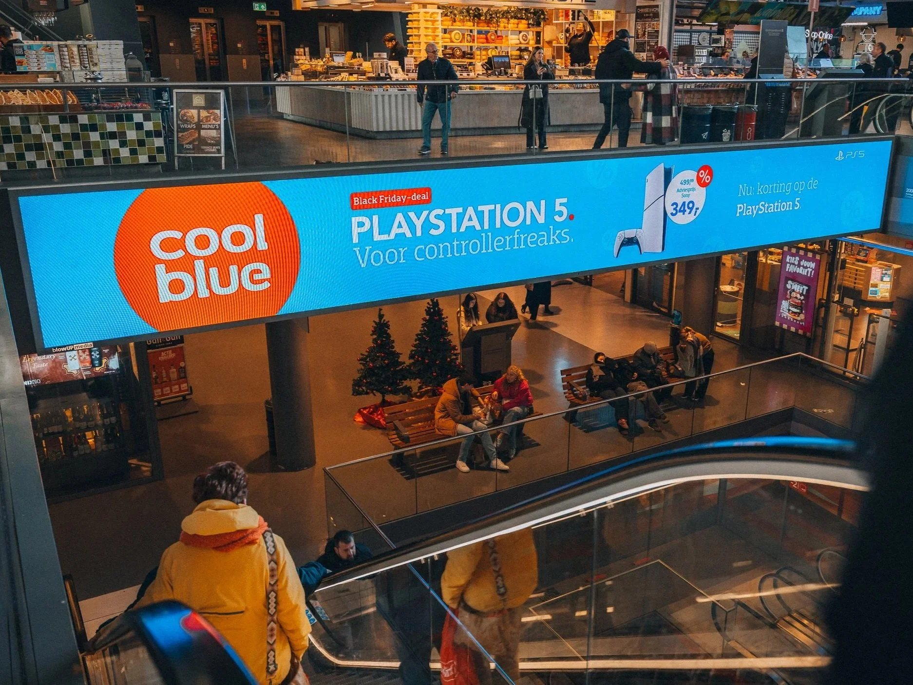 Binnenkant van een winkelcentrum met een grote blauwe reclame banner voor Coolblue met een aanbieding voor PlayStation 5. Mensen wachten, zitten en lopen in het winkelcentrum.