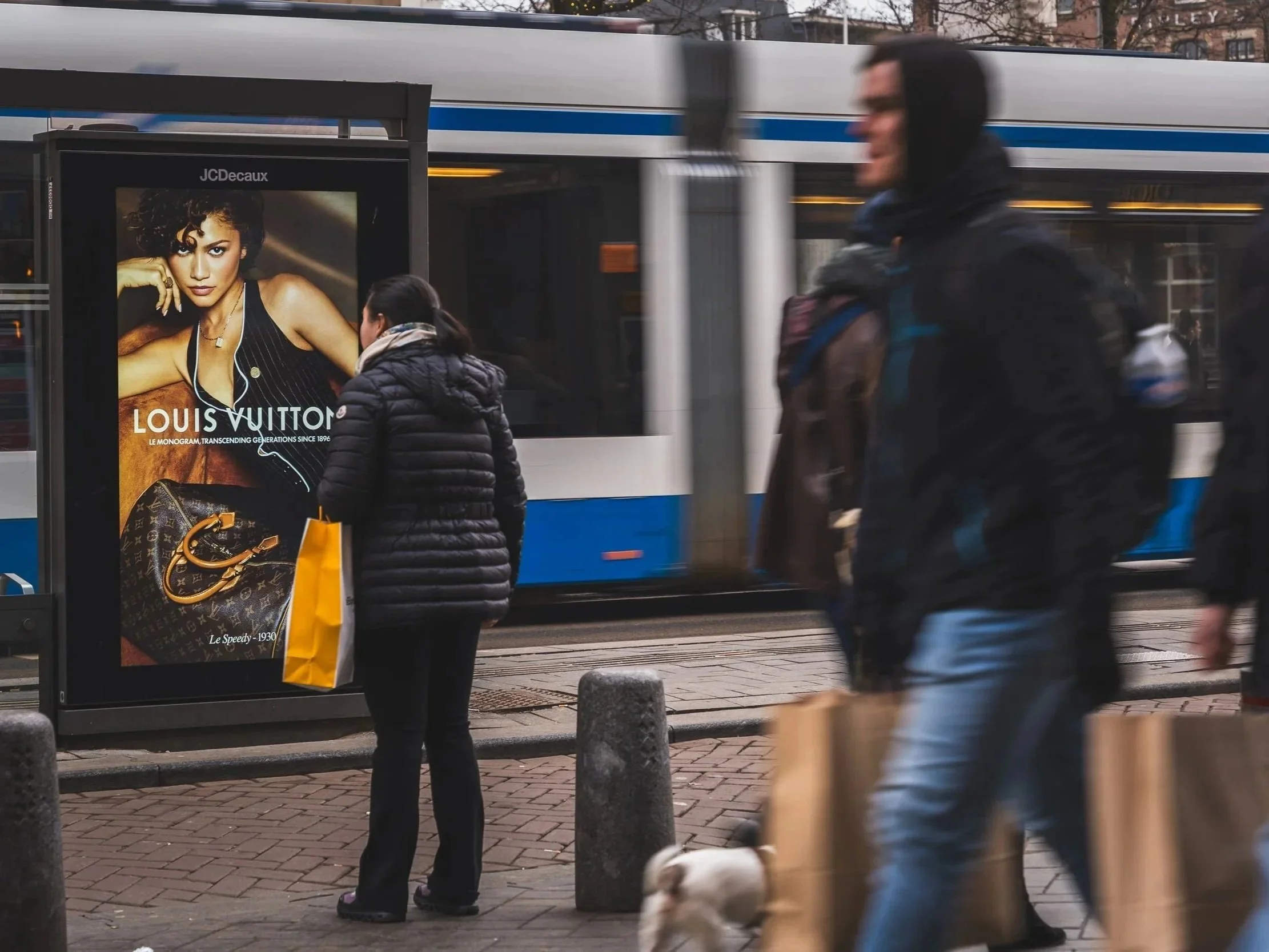 Een vrouw staat voor een Louis Vuitton-advertentie op een bushokje, terwijl een bus voorbij rijdt. Andere mensen lopen langs, één met een hondje en een man met een tas.