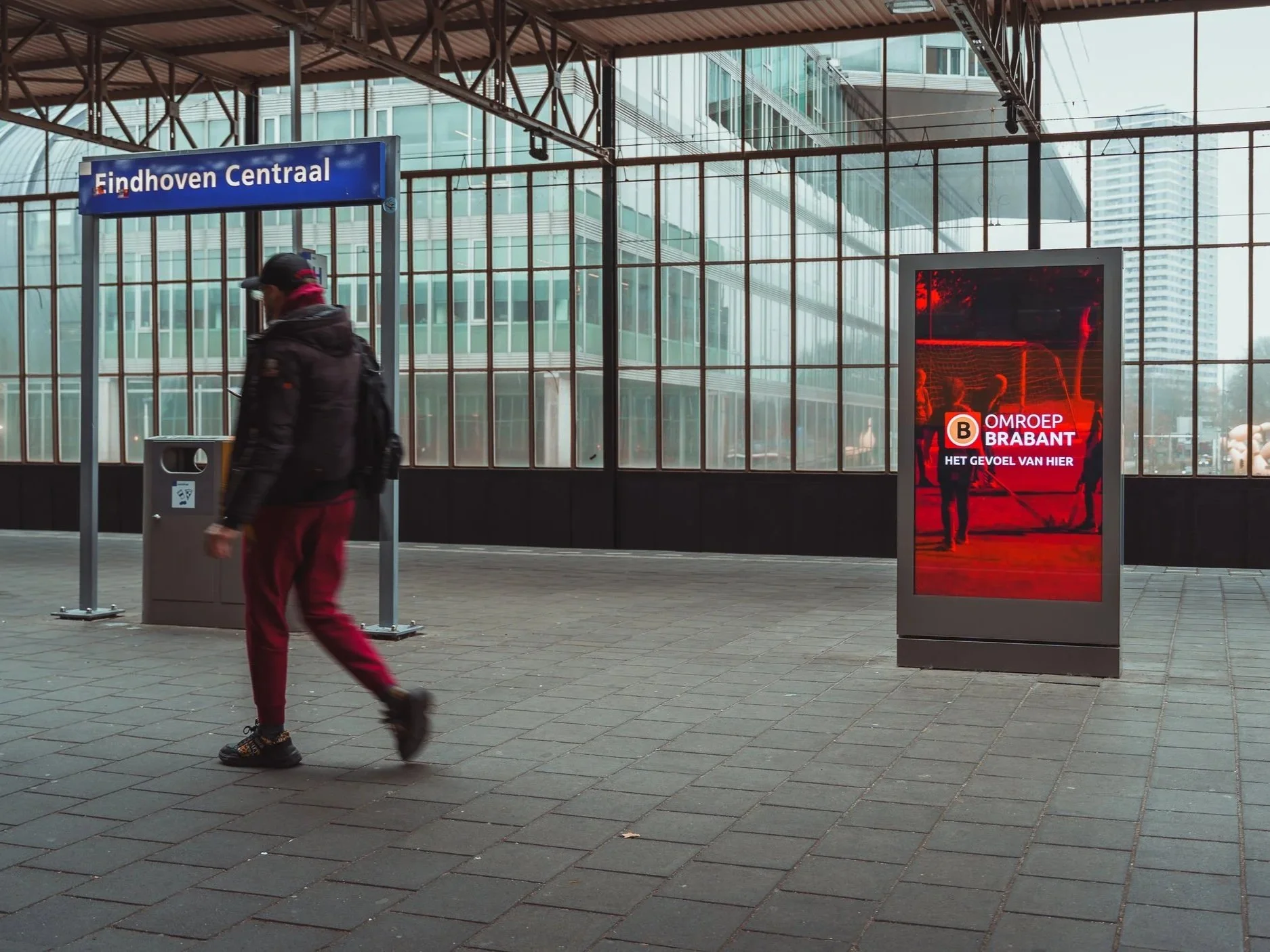 Een persoon loopt door het treinstation Eindhoven Centraal. Er is een digitale informatiezuil en een blauw stationbord met de tekst 'Eindhoven Centraal'. Het station heeft grote glazen wanden met uitzicht op moderne gebouwen.