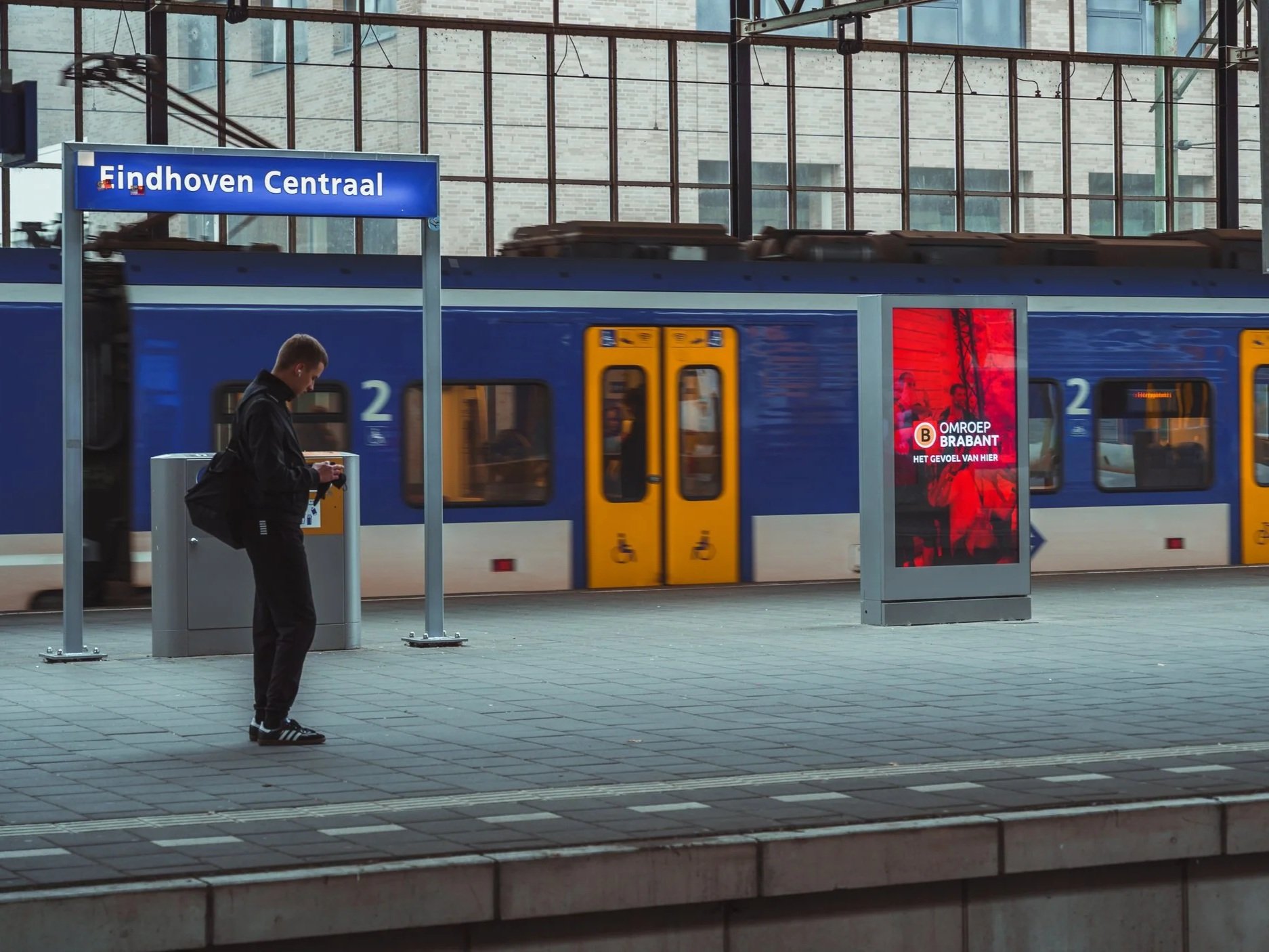 Een persoon wacht op het perron bij Eindhoven Centraal met een trein op de achtergrond, een digitale reclamezuil en een blauw en geel treinstationbord.