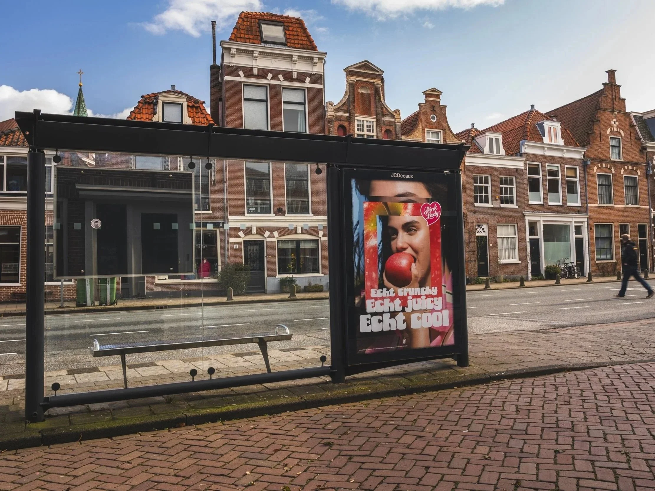 Straatbeeld met een bushokje en een reclamebord. De achtergrond bestaat uit historische bakstenen gebouwen met zadeldaken. In de straat lopen mensen en staan fietsen.