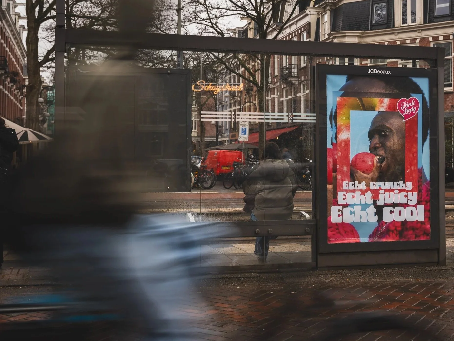 Een bushokje met een poster van een persoon die een rode fruit vasthoudt, met teksten in het Nederlands die zeggen: "Echt crunchy, echt juicy, echt cool."
