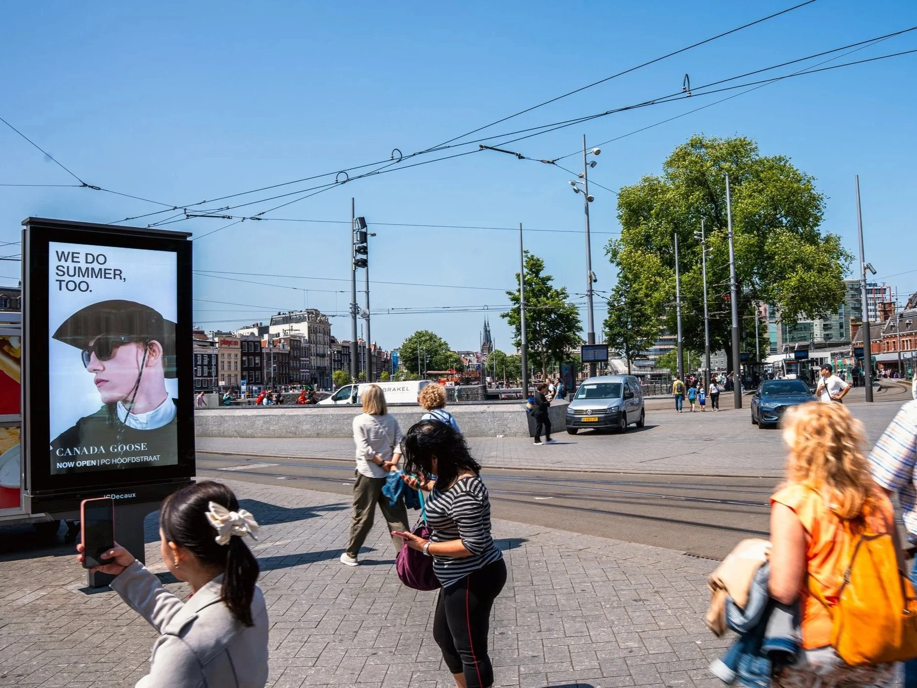 Stadsplein met mensen, auto’s, tramrails, reclamebord met een persoon met hoed en zonnebril, bomen en gebouwen, onder een blauwe hemel.