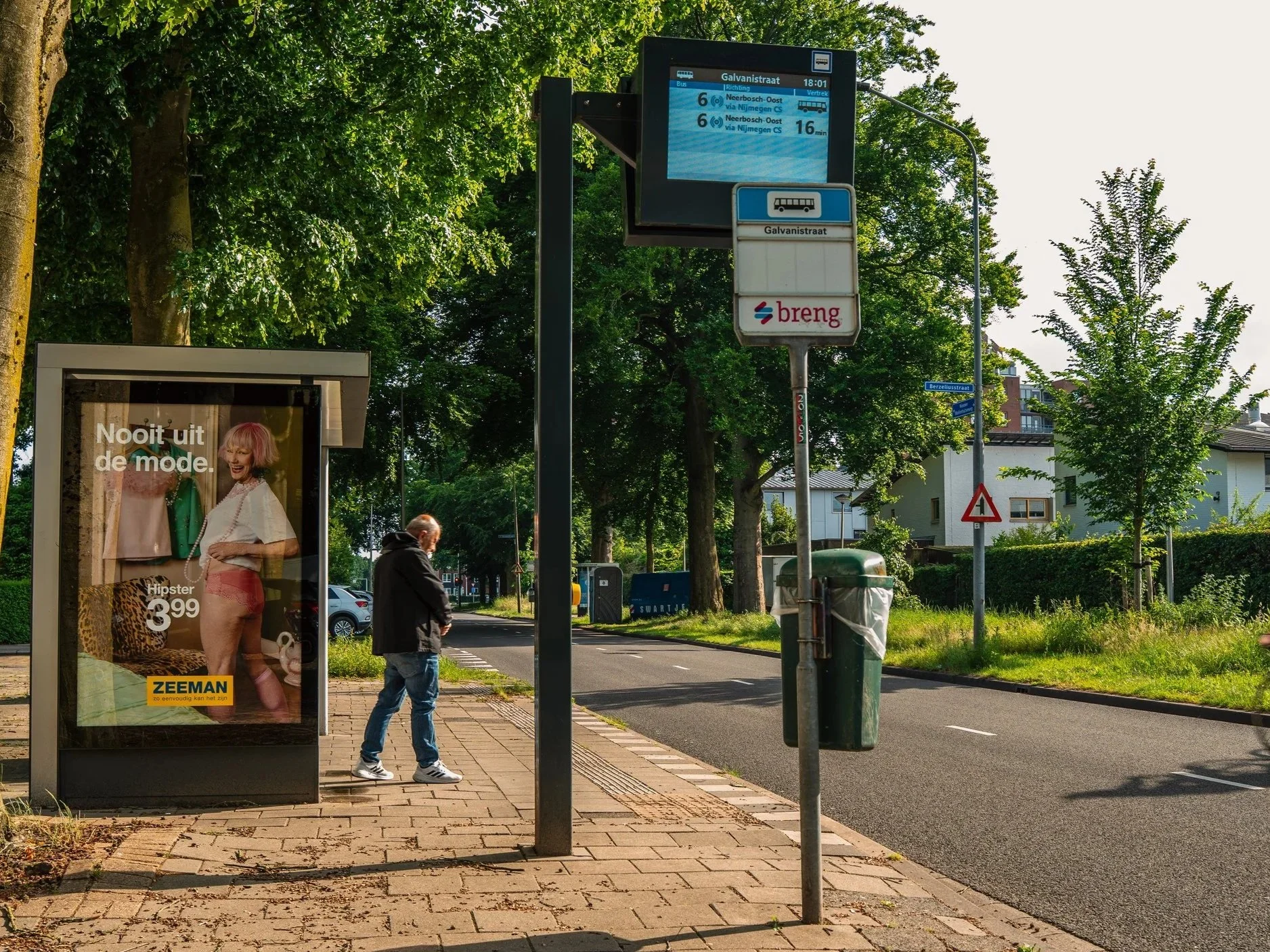 Een bushokje met een advertentie, een persoon die staat te wachten, een bushalte bord en een digitale vertrektijden display op een zonnige straat met bomen en huizen in de achtergrond.