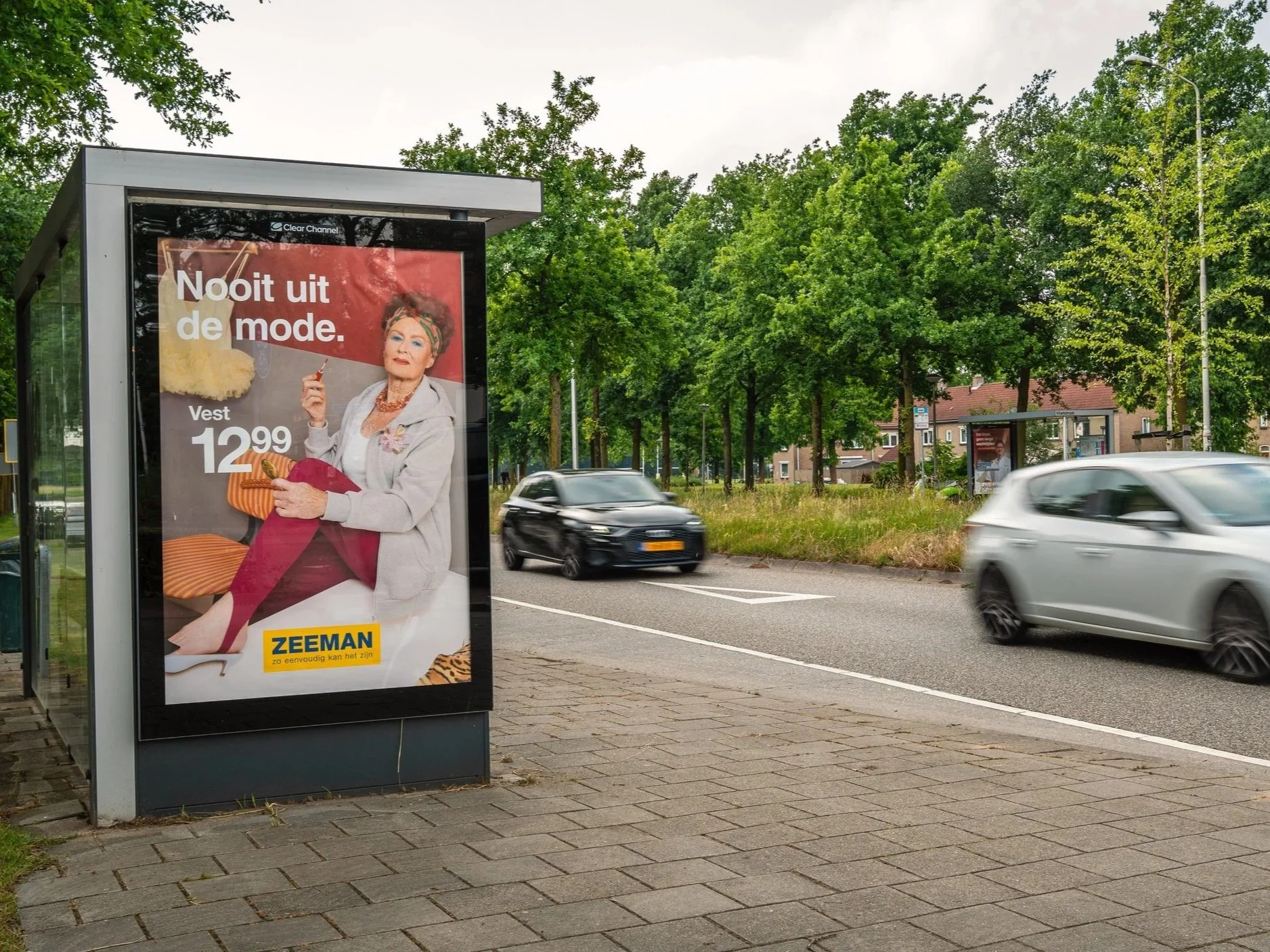 Advertentie met een oudere vrouw die in een witte jas zit en rode broek, op een bankje. Tekst op de advertentie zegt 'Nooit uit de mode. Vest 12.99'.