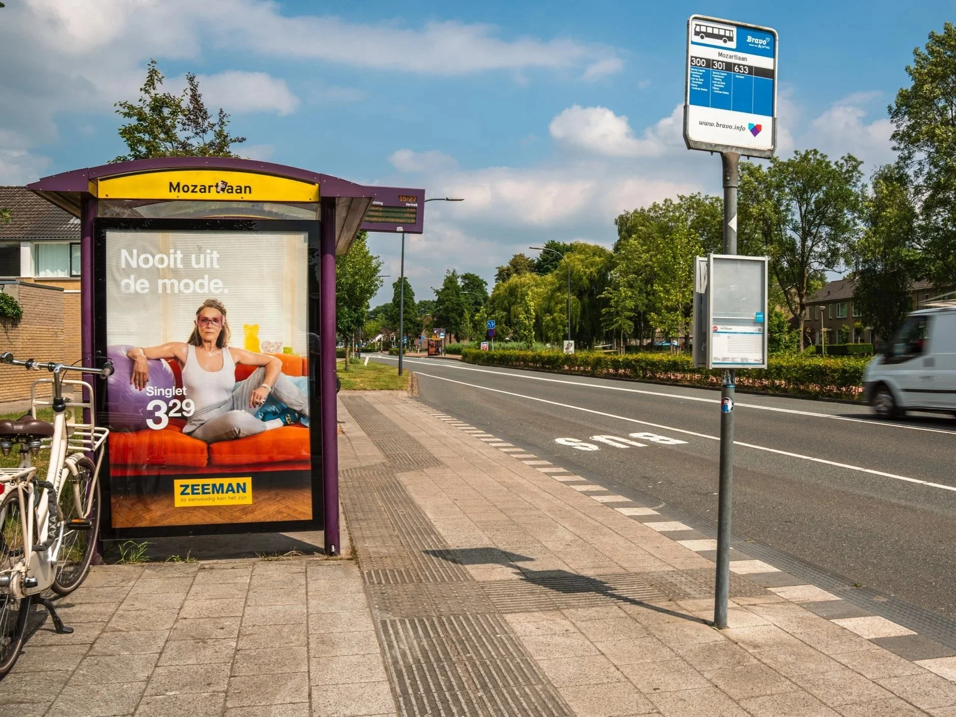 Een bushokje met een advertentie voor Zeeman, een reclame met een vrouw die ontspannen op een bank zit, en het fietspad, de weg en een busstationbord. De omgeving bestaat uit bomen en woningen.
