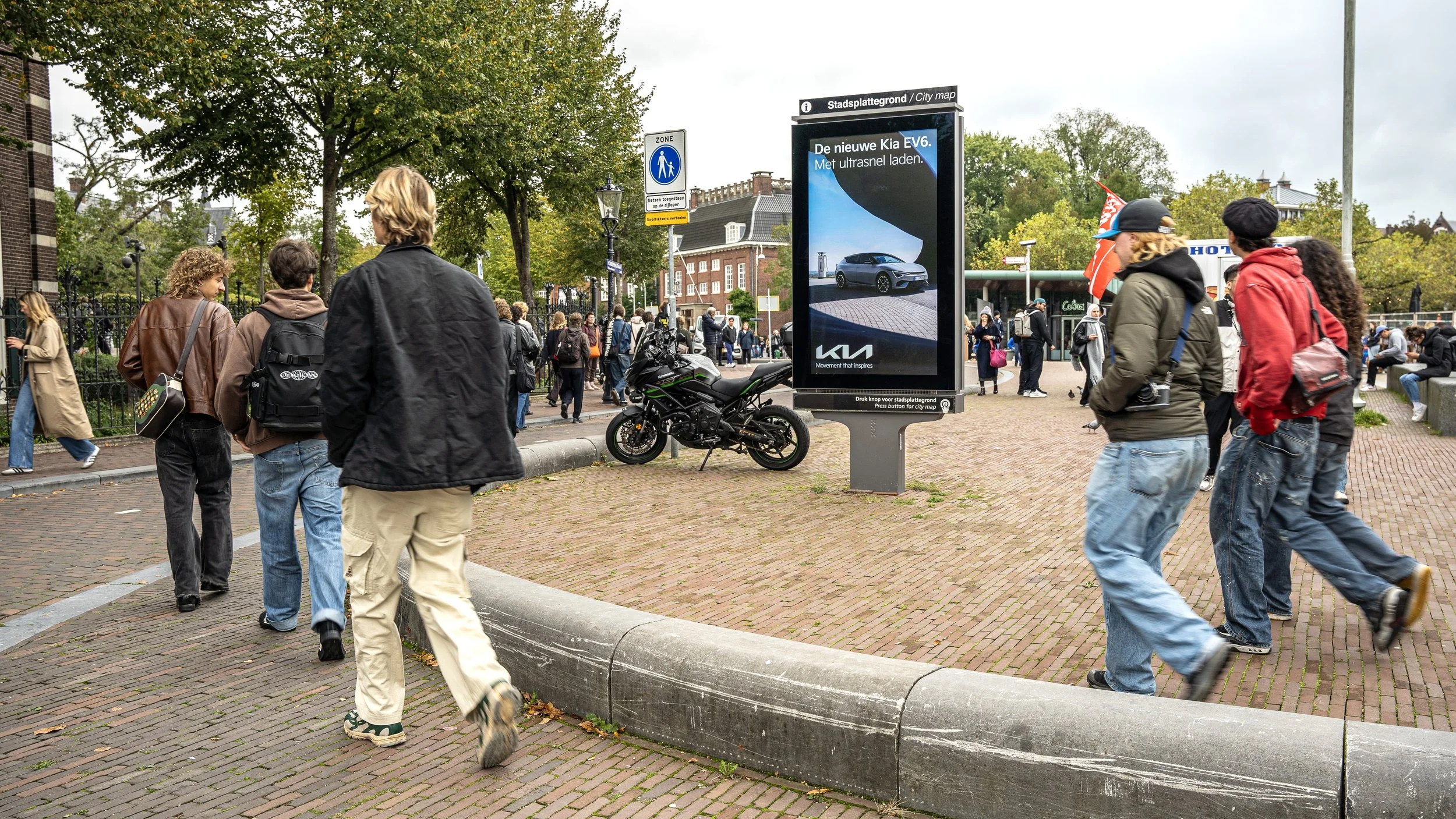 Veel mensen lopen op een straat met een digitale reclamezuil en een stilstaande motorfiets. In de achtergrond zijn bomen en gebouwen zichtbaar.