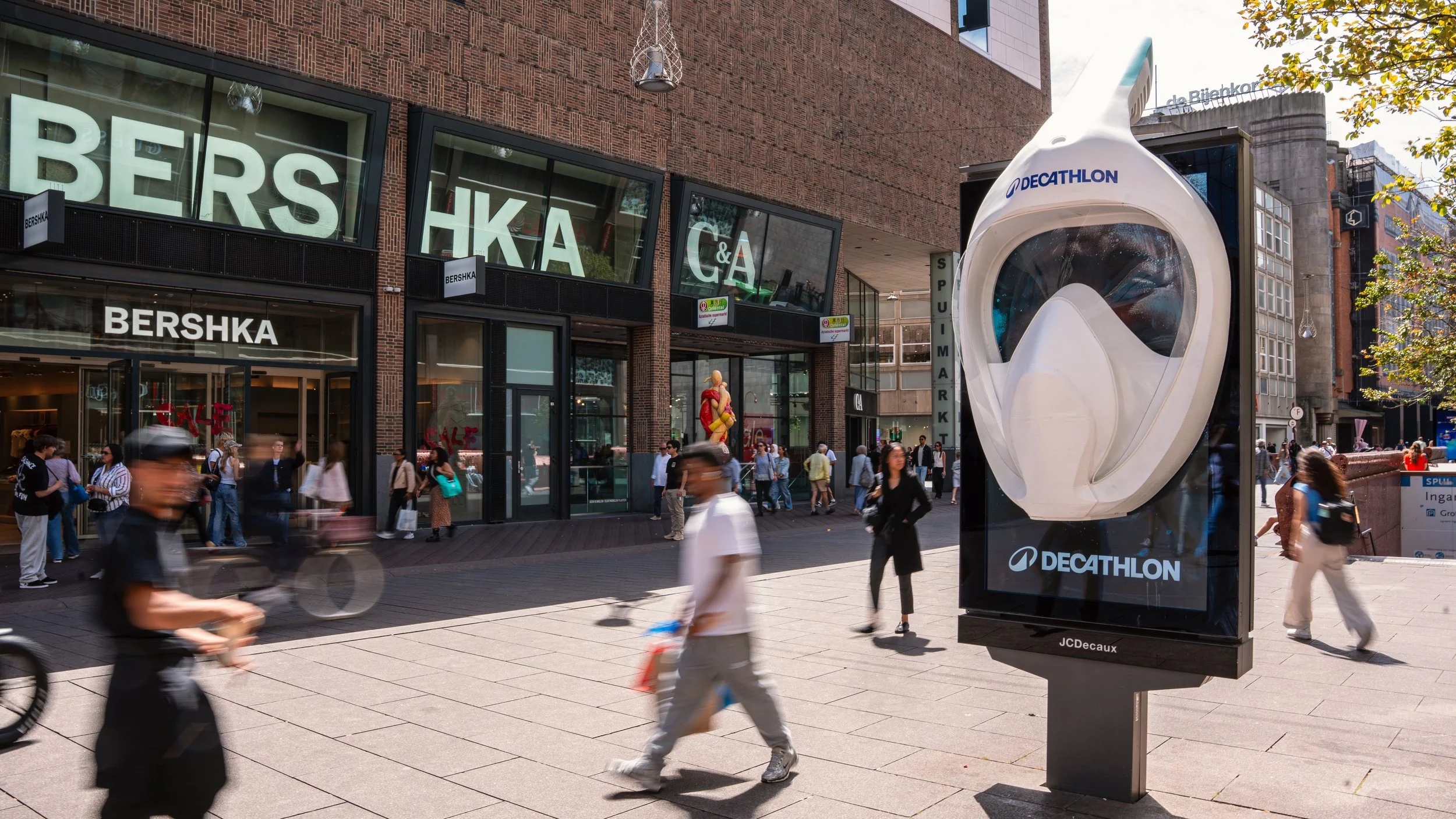 Straatbeeld met mensen die lopen en fietsen voor een winkelcentrum met grote ramen, een poster en een borstmasker van Decathlon.