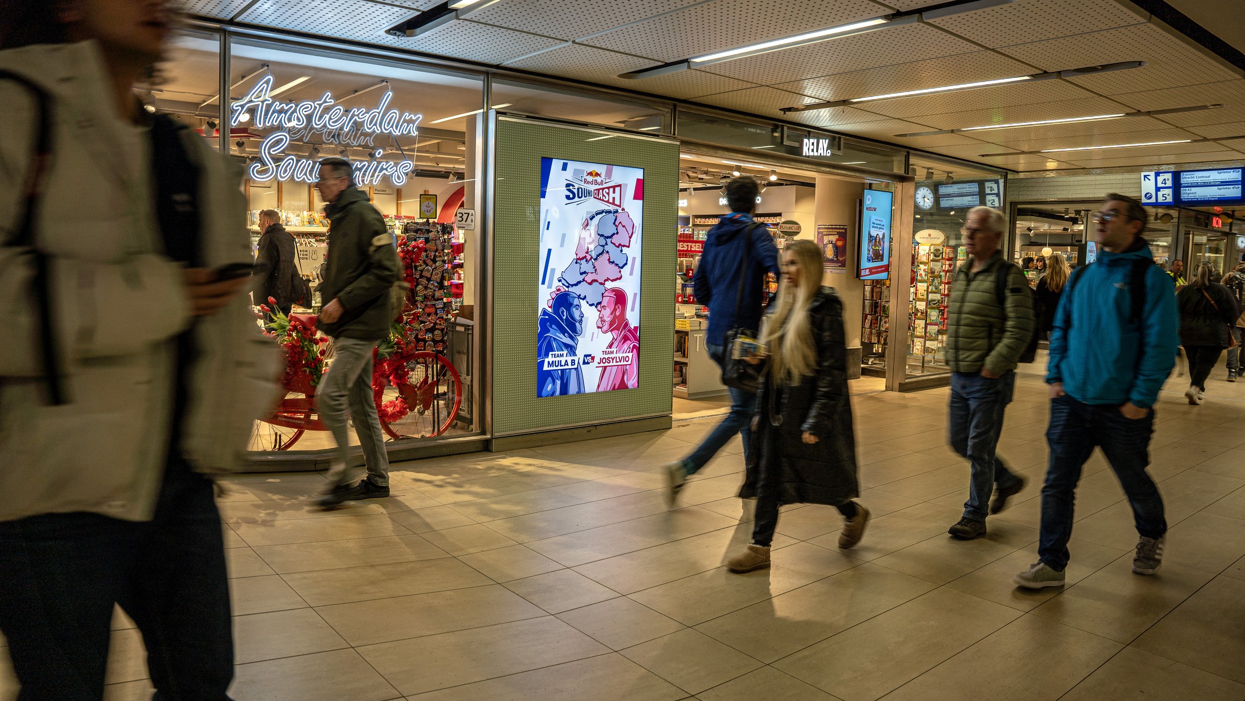 Binnenstad met mensen die lopen, een winkel met opvallende reclame en digitale schermen, en een digitale posters met sportevenement.