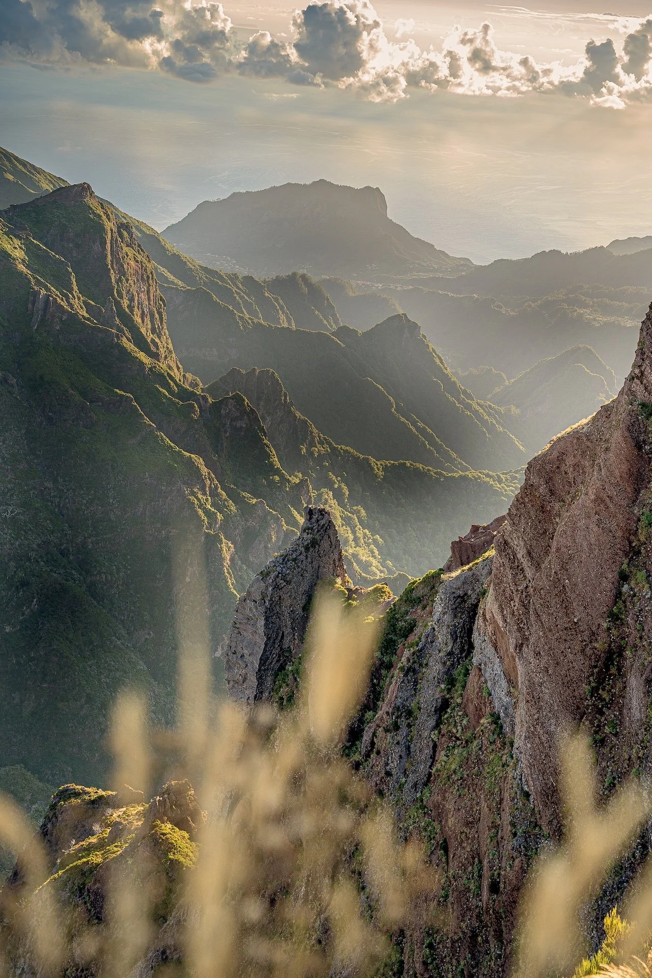 Glooiende groene bergen met rotsformaties en een zachte mist die over de valleien hangt, onder een bewolkte hemel met zonnestralen.