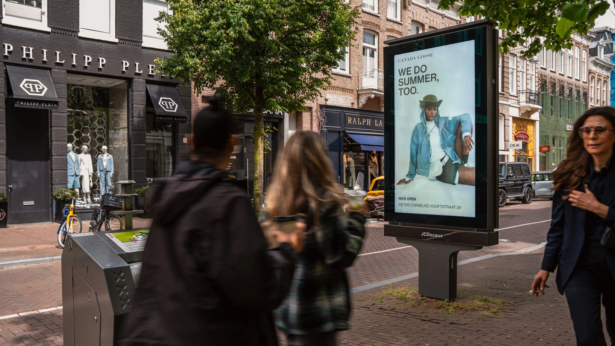 Een straat in Amsterdam met winkels en voetgangers, een digitale reclamezuil met een model en tekst in het Engels en Nederlands, en een fiets.
